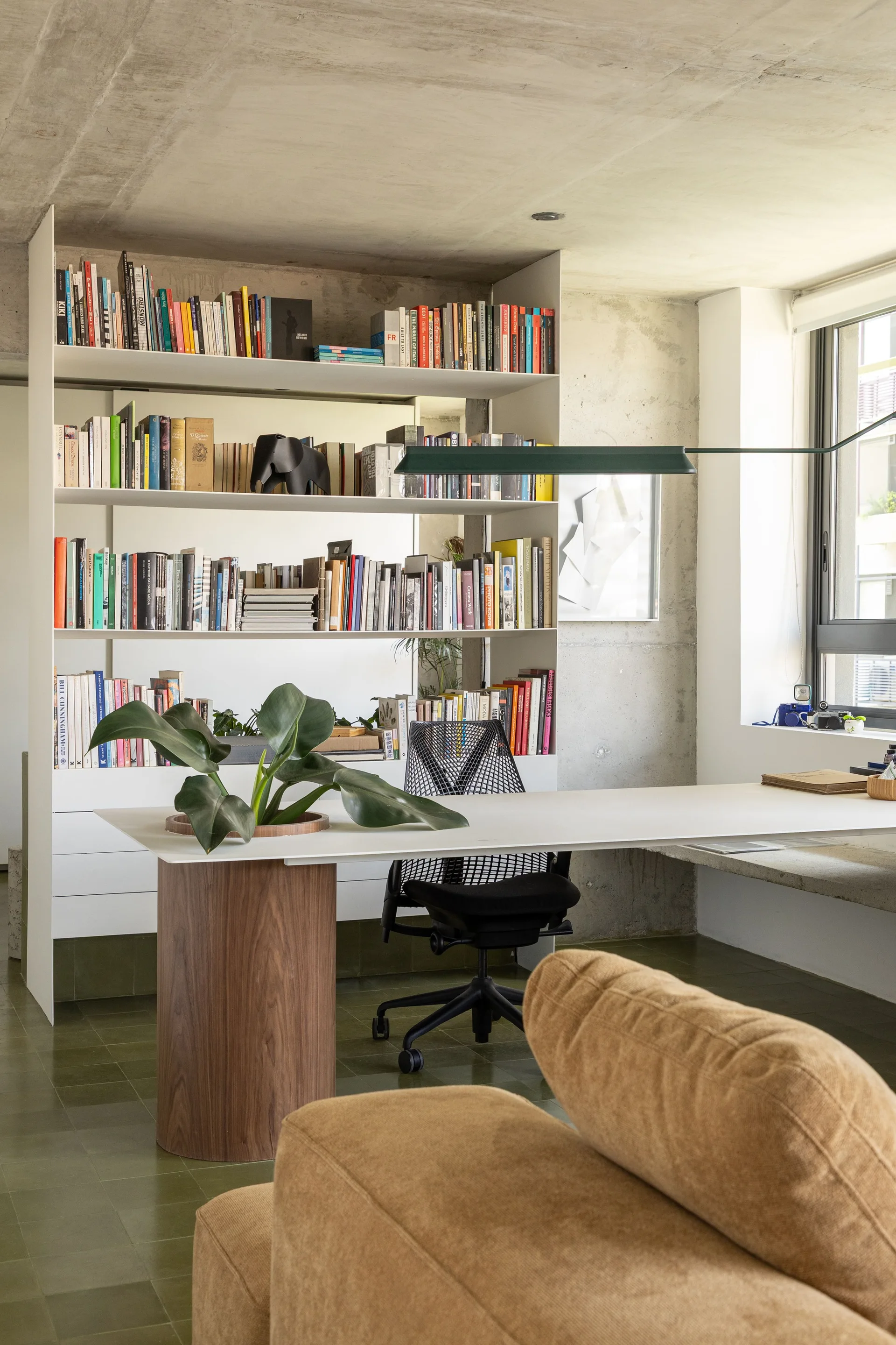 A modern home office featuring a white desk with a cylindrical wood base, a tall white bookshelf, and exposed raw concrete walls and ceiling.