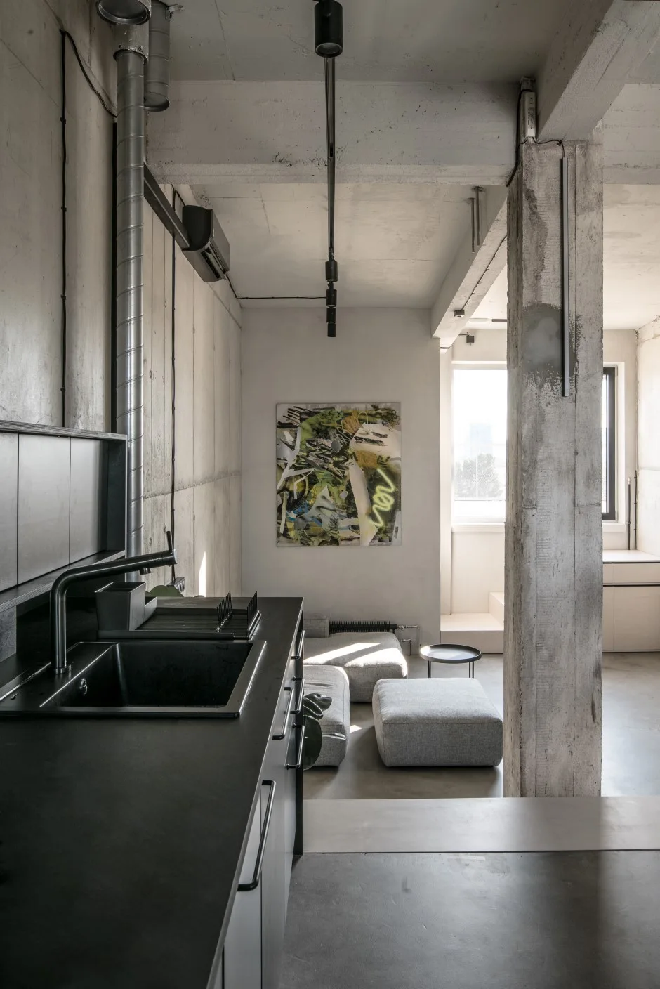 An industrial loft features a matte black kitchen counter in the foreground, exposed concrete walls, and low-profile gray seating in the background.