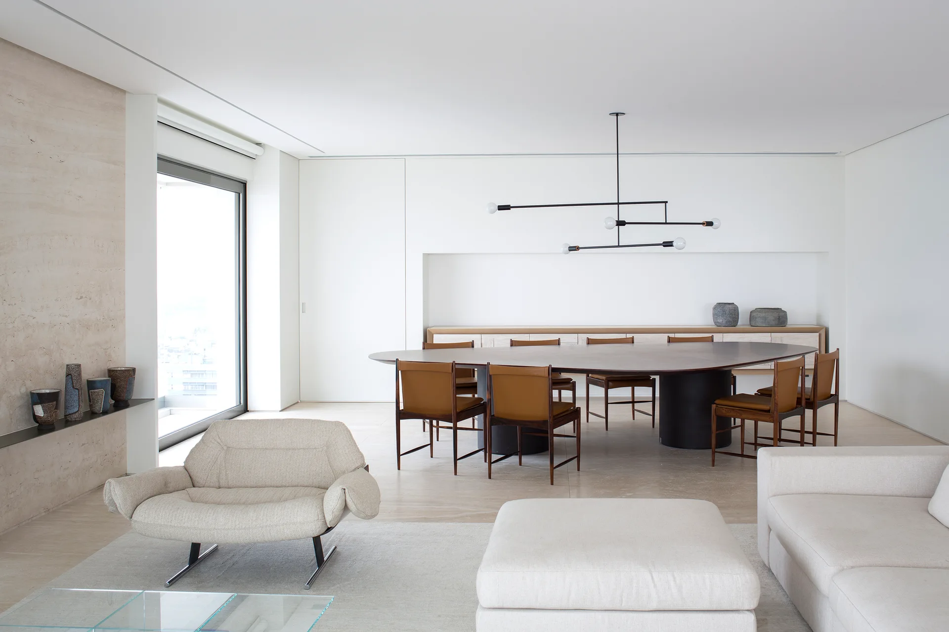A minimalist open-plan dining area features a large oval wood table surrounded by tan leather chairs under a black linear chandelier.