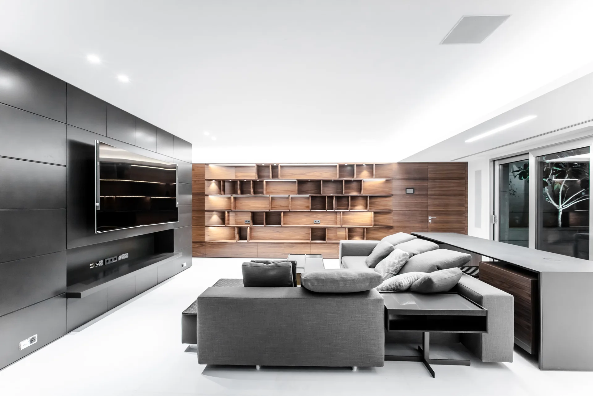A modern living room features a gray modular sectional sofa positioned between a matte black media wall and wood-grained built-in shelving.