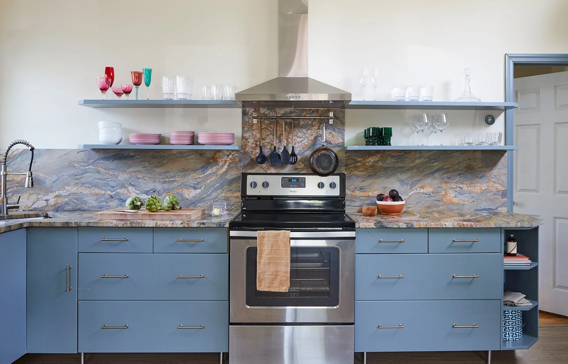 A kitchen featuring matte blue lower cabinets, a highly veined stone countertop and backsplash, a stainless steel range, and matching blue open shelves.