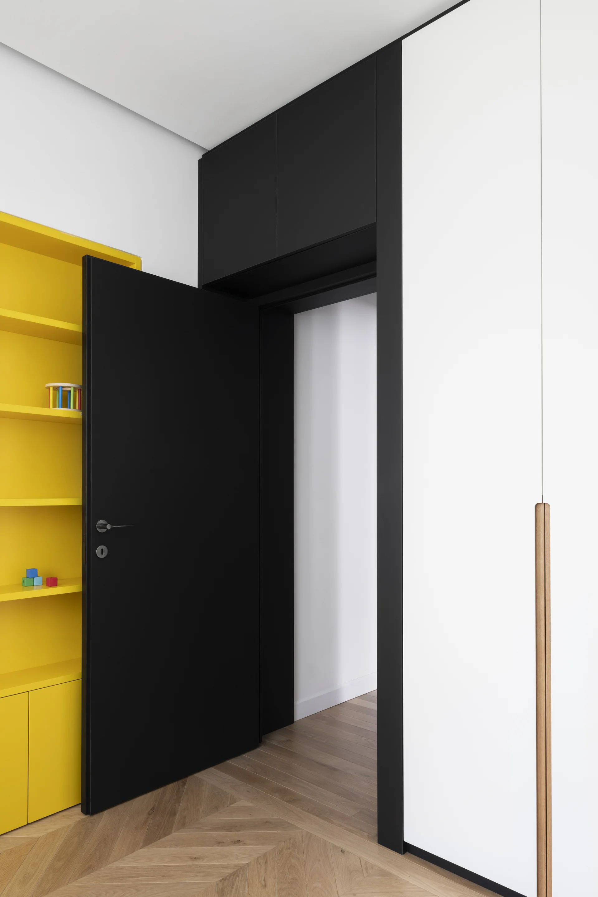 A matte black door stands open next to bright yellow open shelving and a tall white wardrobe on light wood herringbone flooring.