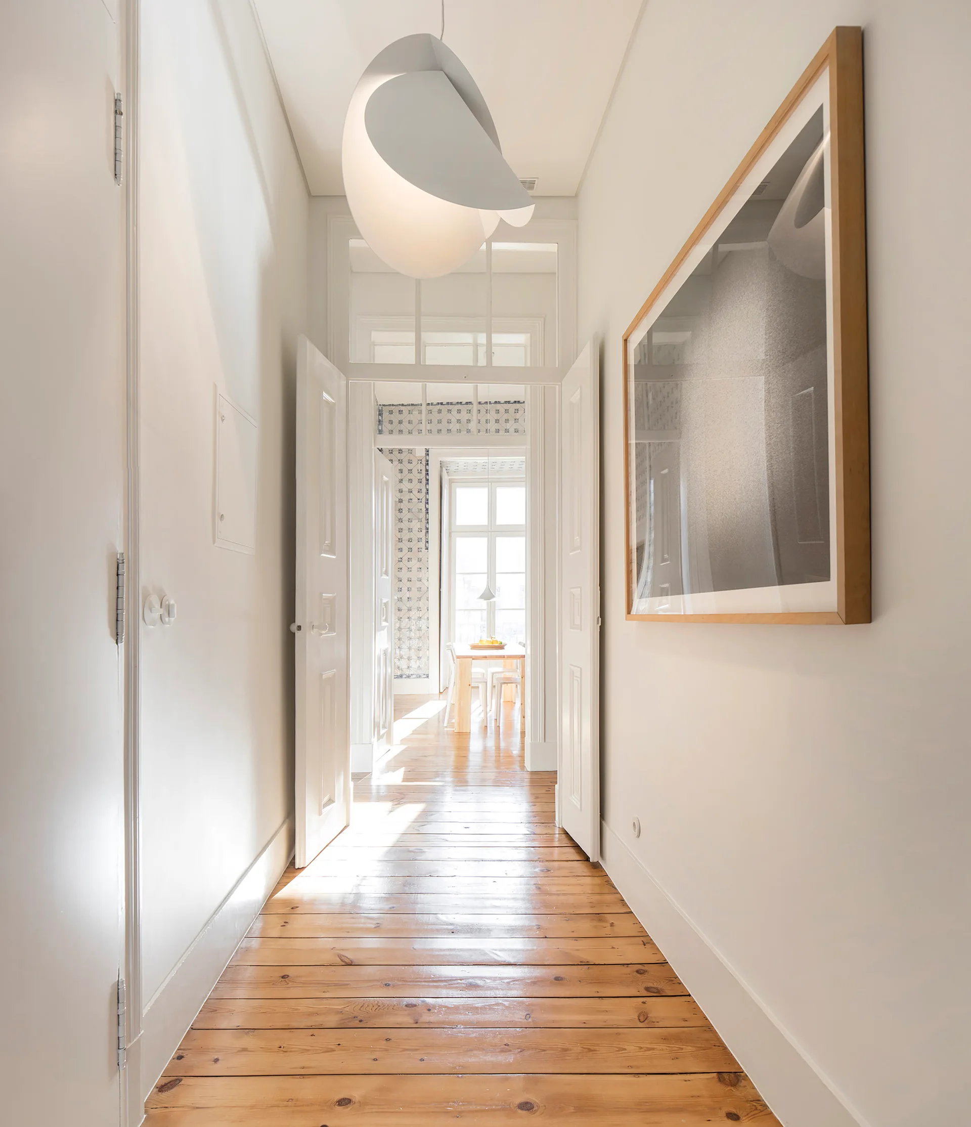 A bright hallway with glossy wood plank floors features white walls, open double doors, a transom window, and a sculptural pendant light.