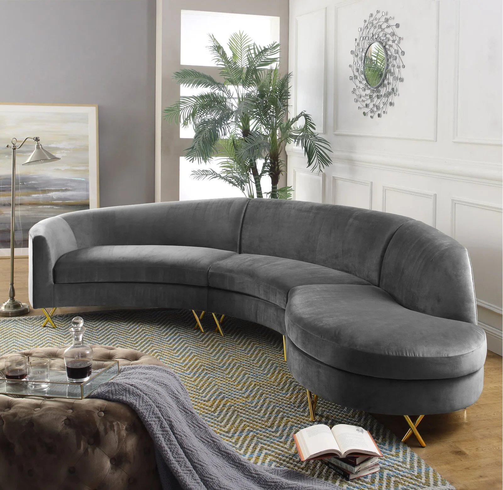 A curved grey velvet sectional sofa sits on a chevron patterned rug in a room with wainscoted walls and a round mirror.