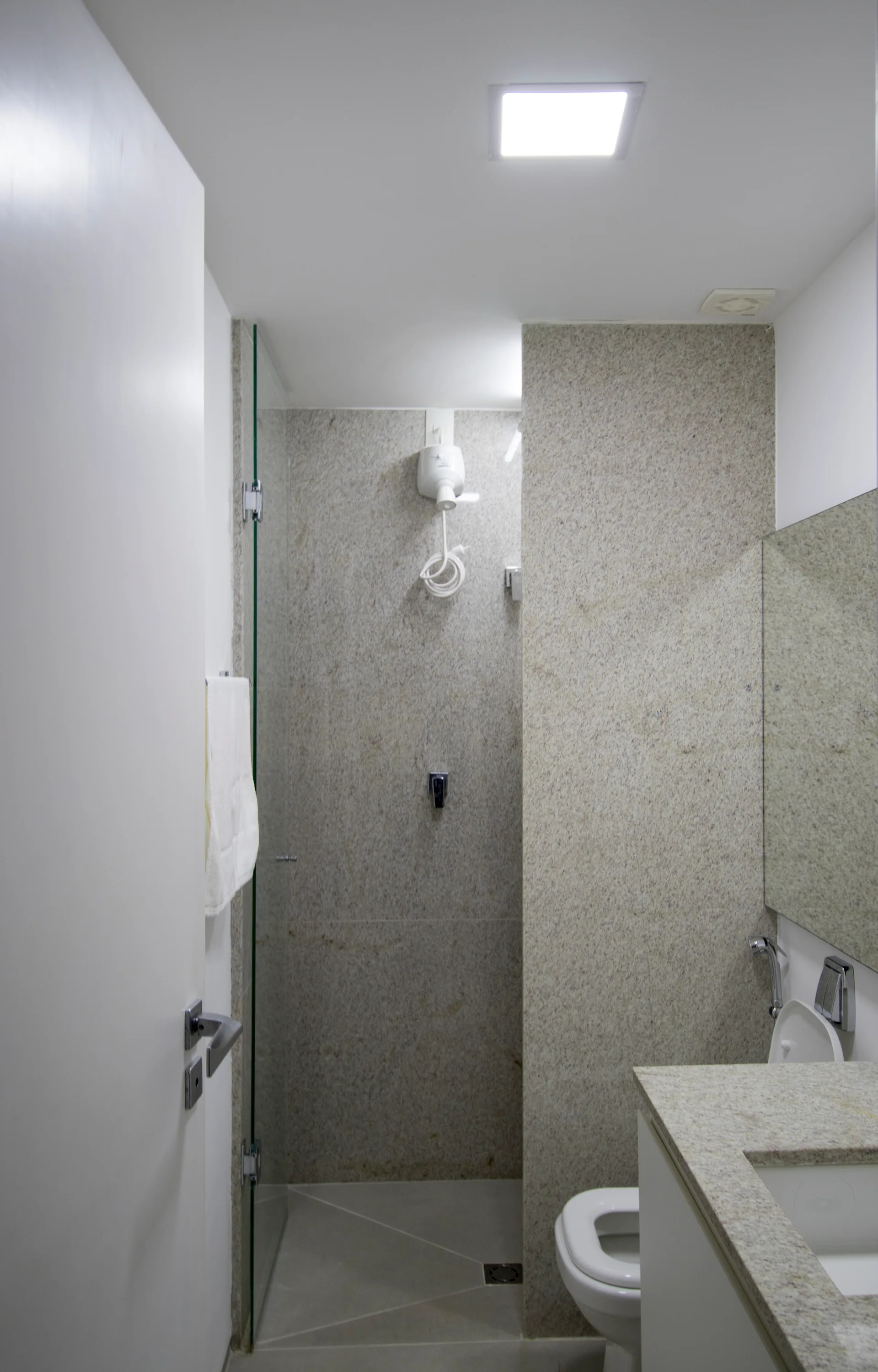 A small modern bathroom featuring beige speckled stone walls, a glass shower door, a white toilet, and a matching stone vanity countertop.