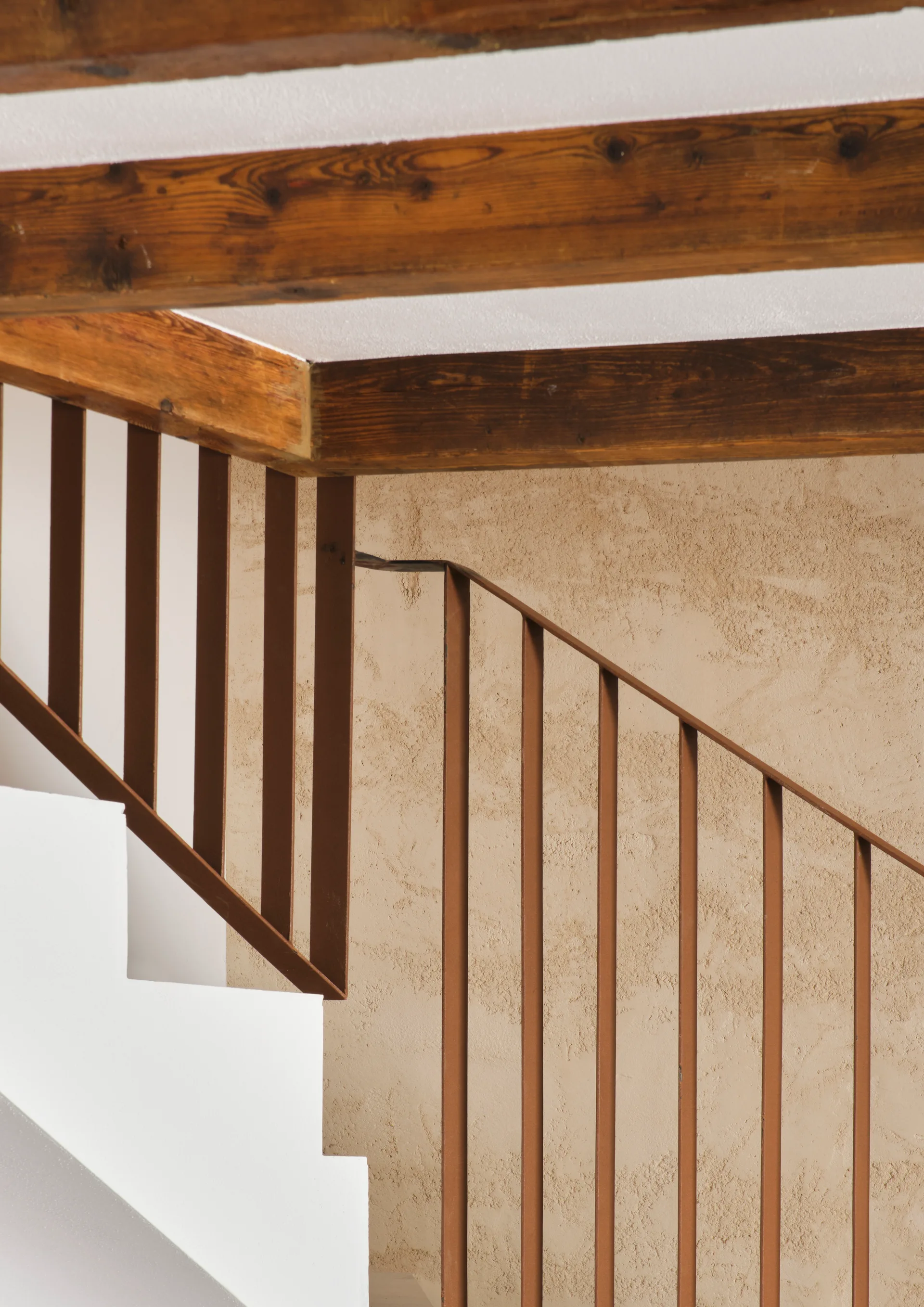 A white staircase with a slender brown metal railing sits against a textured beige plaster wall beneath exposed wooden ceiling beams.