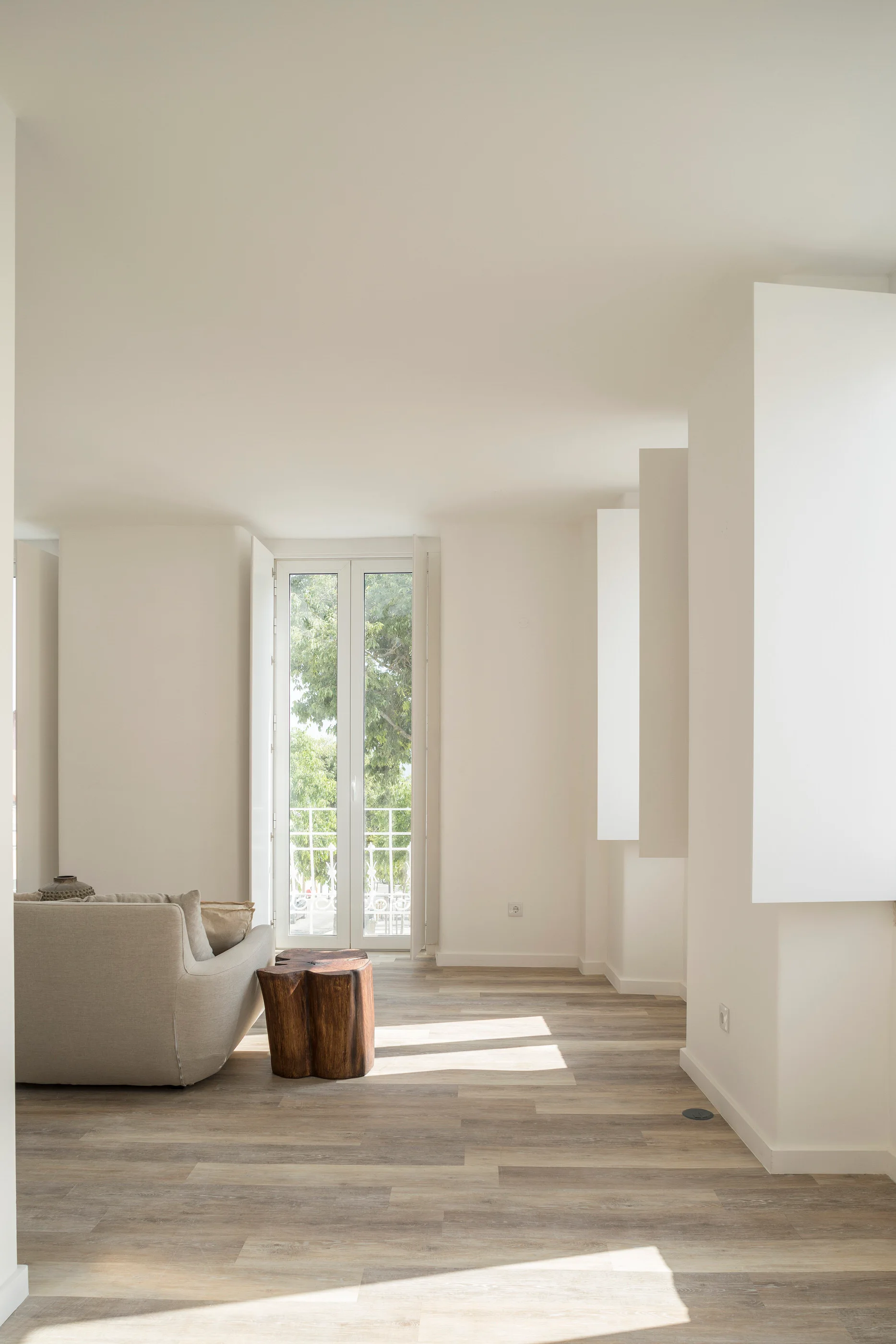 A minimalist living room features a beige sofa and a polished wood stump side table on light-colored plank flooring next to white walls.