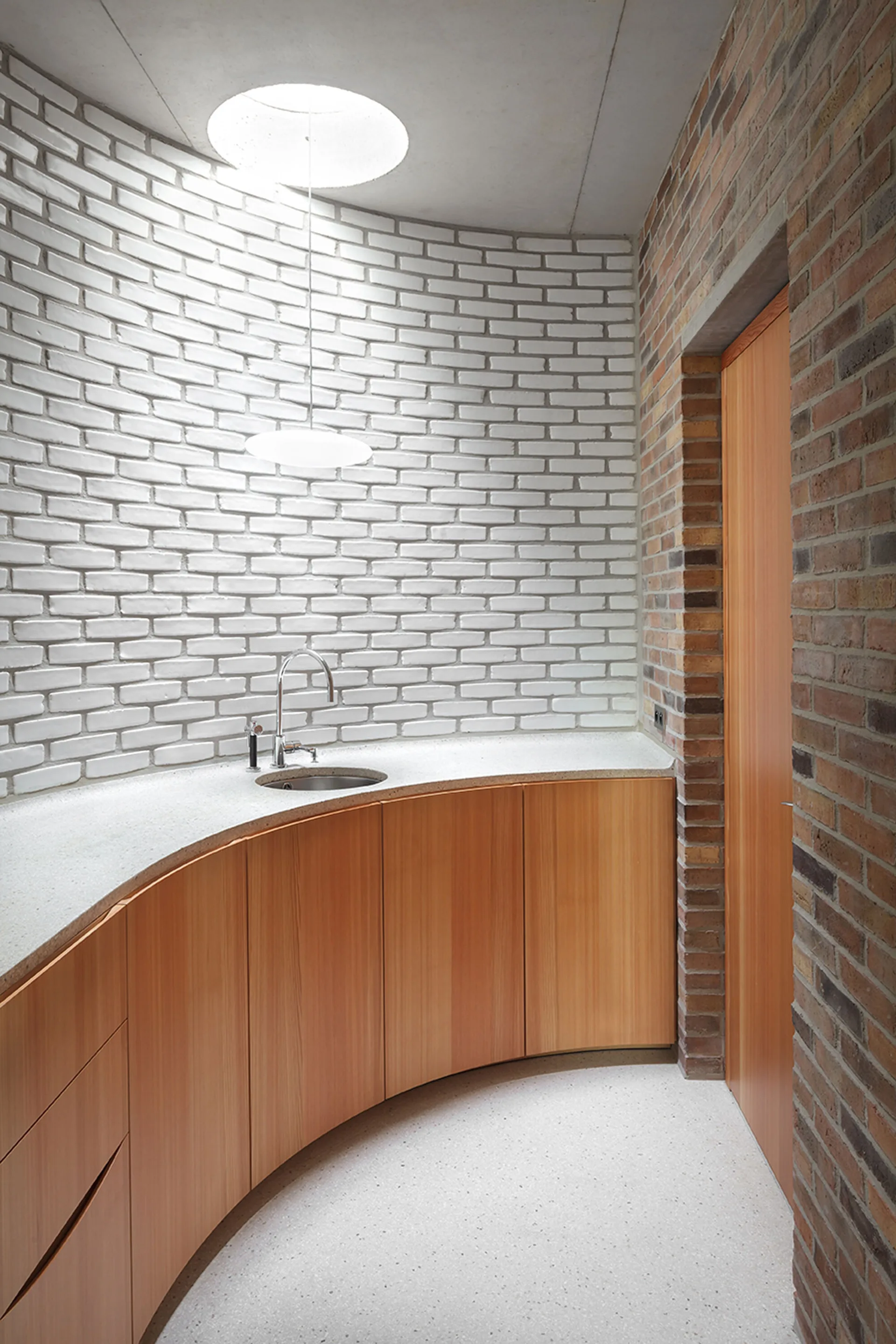 A curved wooden vanity with a metal sink is set against a white-painted brick wall below a circular skylight and minimalist pendant light.