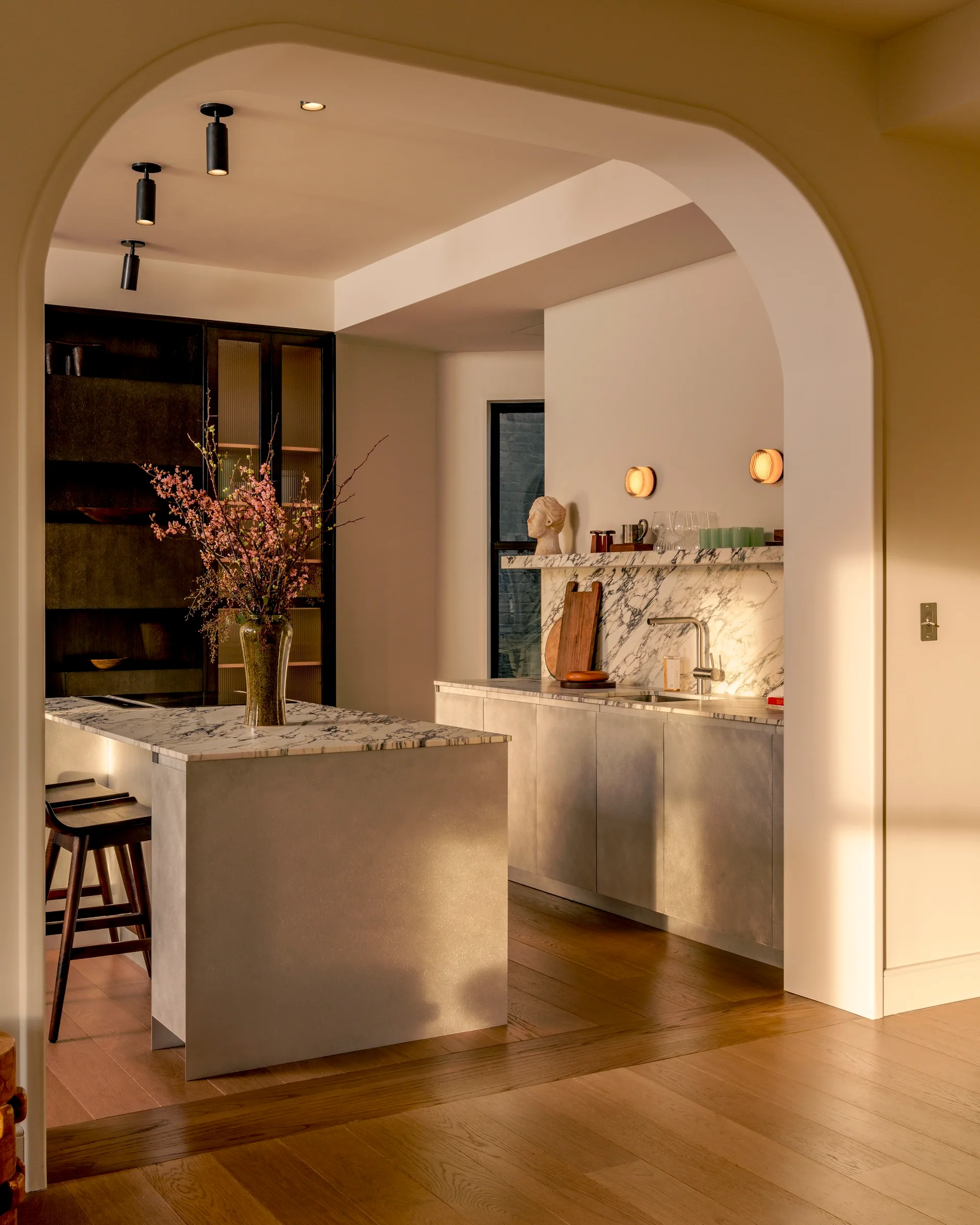 A minimalist kitchen featuring a marble island and matching countertops is viewed through a large plastered archway.
