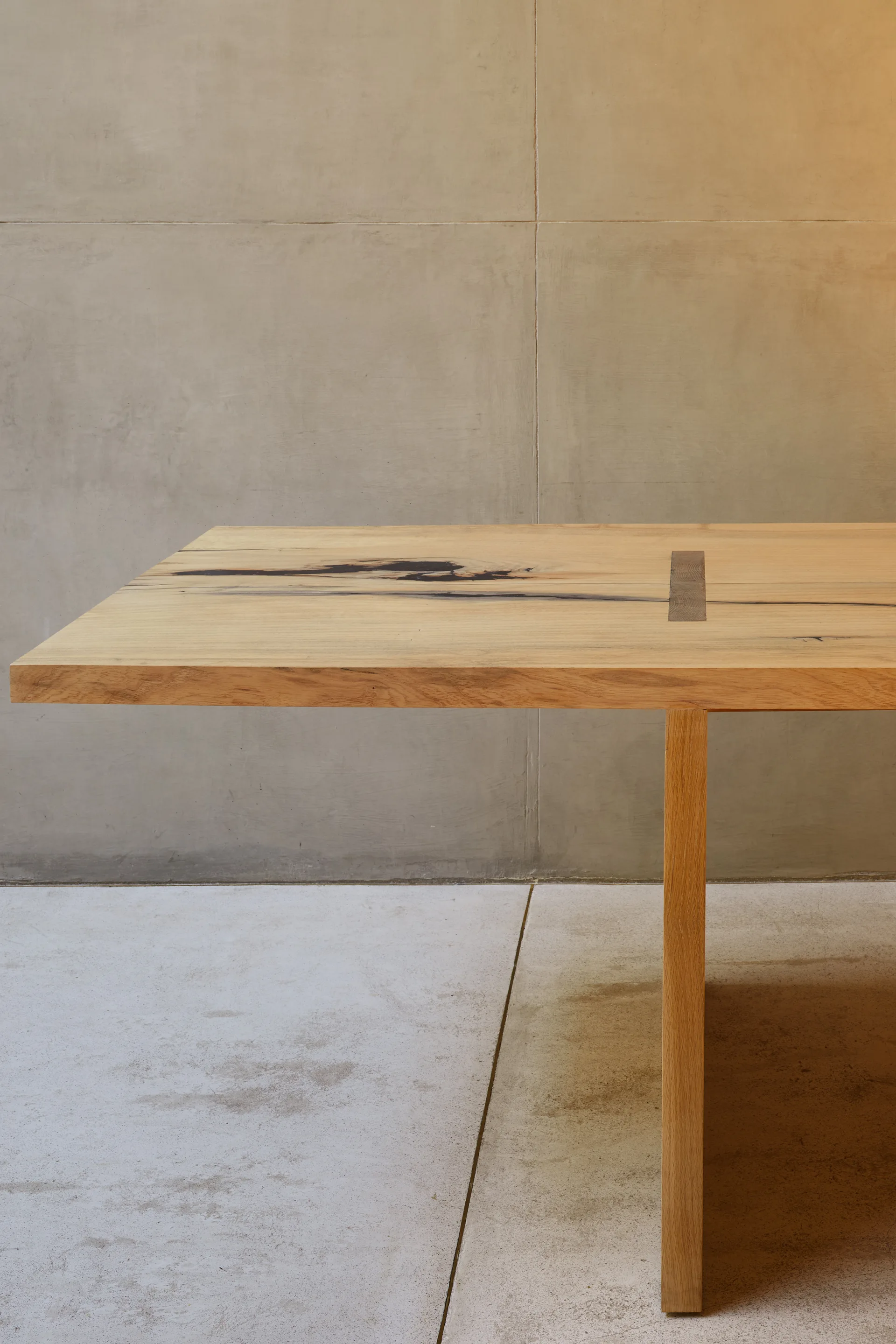 A light wood dining table with a dark rectangular inlay detail stands against a smooth grey concrete paneled wall.