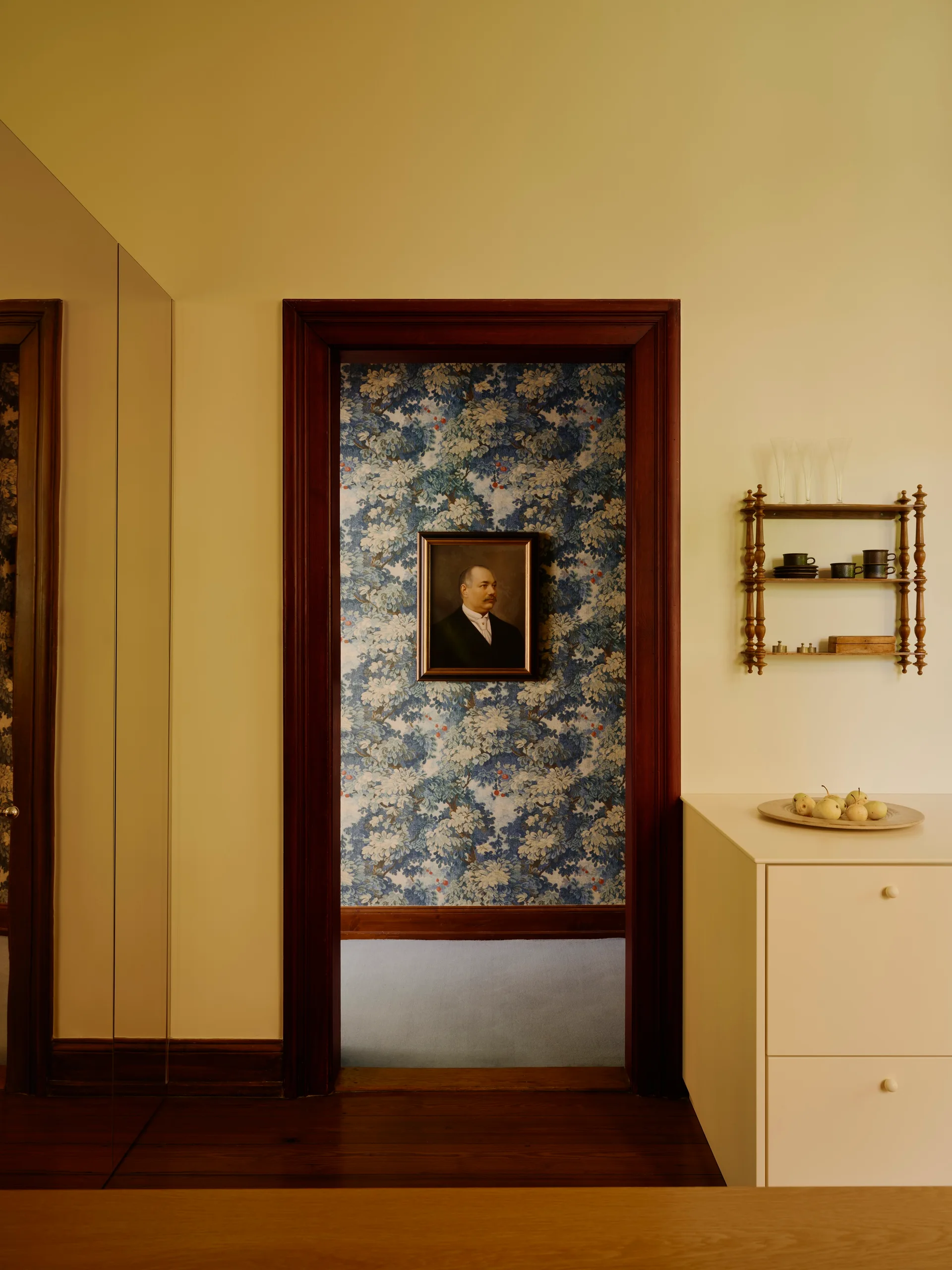 A view through a dark wood doorway shows a room with blue floral wallpaper and a framed portrait, next to a cream cabinet and wooden wall shelf.