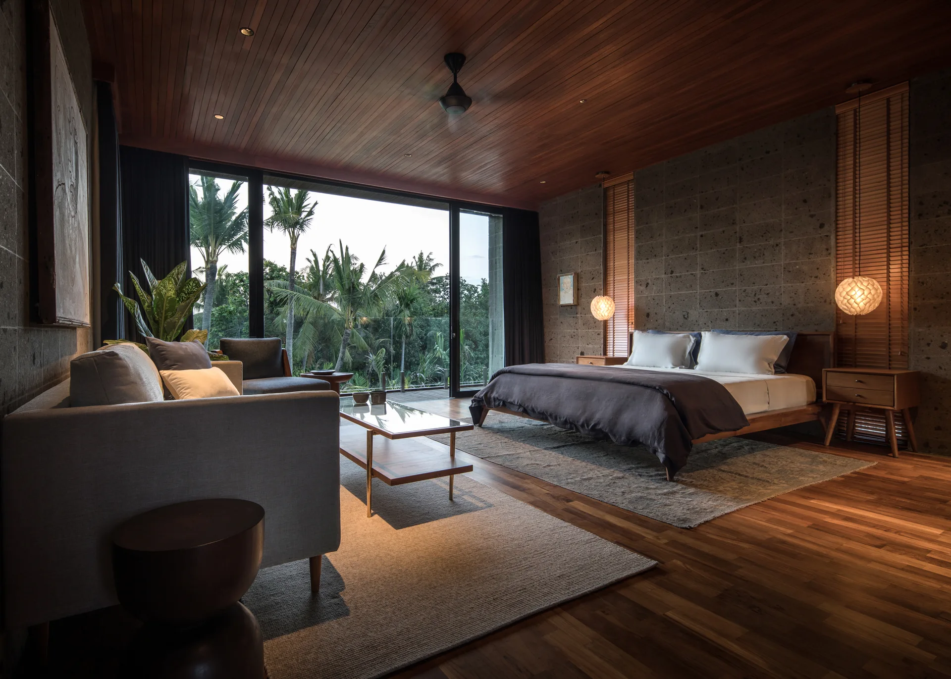 A modern tropical bedroom featuring a dark stone wall, wood plank ceiling, and sliding glass doors that open to lush green trees.