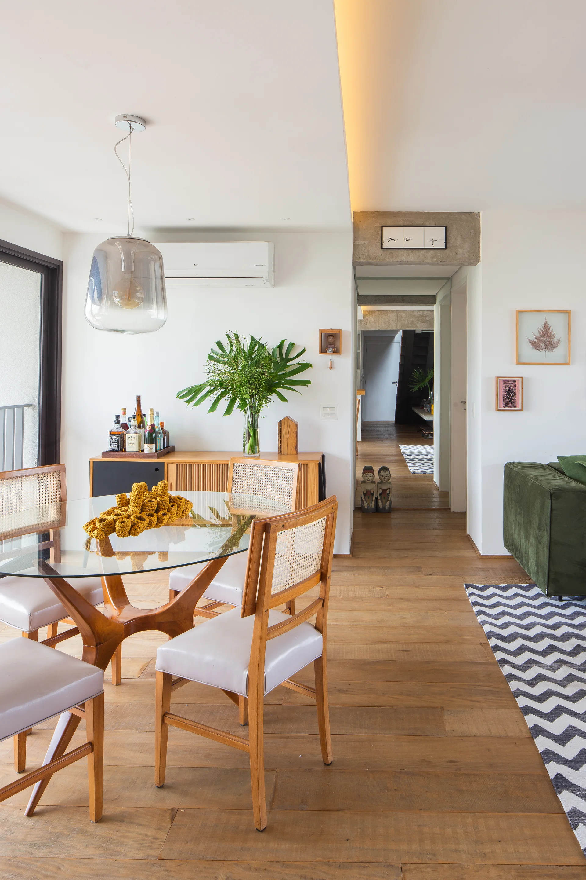 A bright dining room features a round glass-topped table with cane back chairs, a wood sideboard, and a view into a hallway.