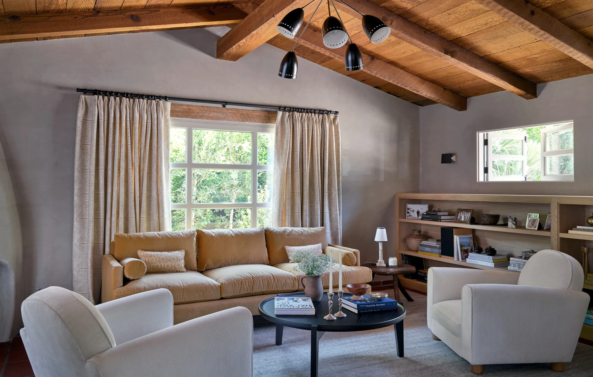 A cozy living room features a mustard velvet sofa, two cream armchairs, and a black coffee table beneath a vaulted wood ceiling with exposed beams.