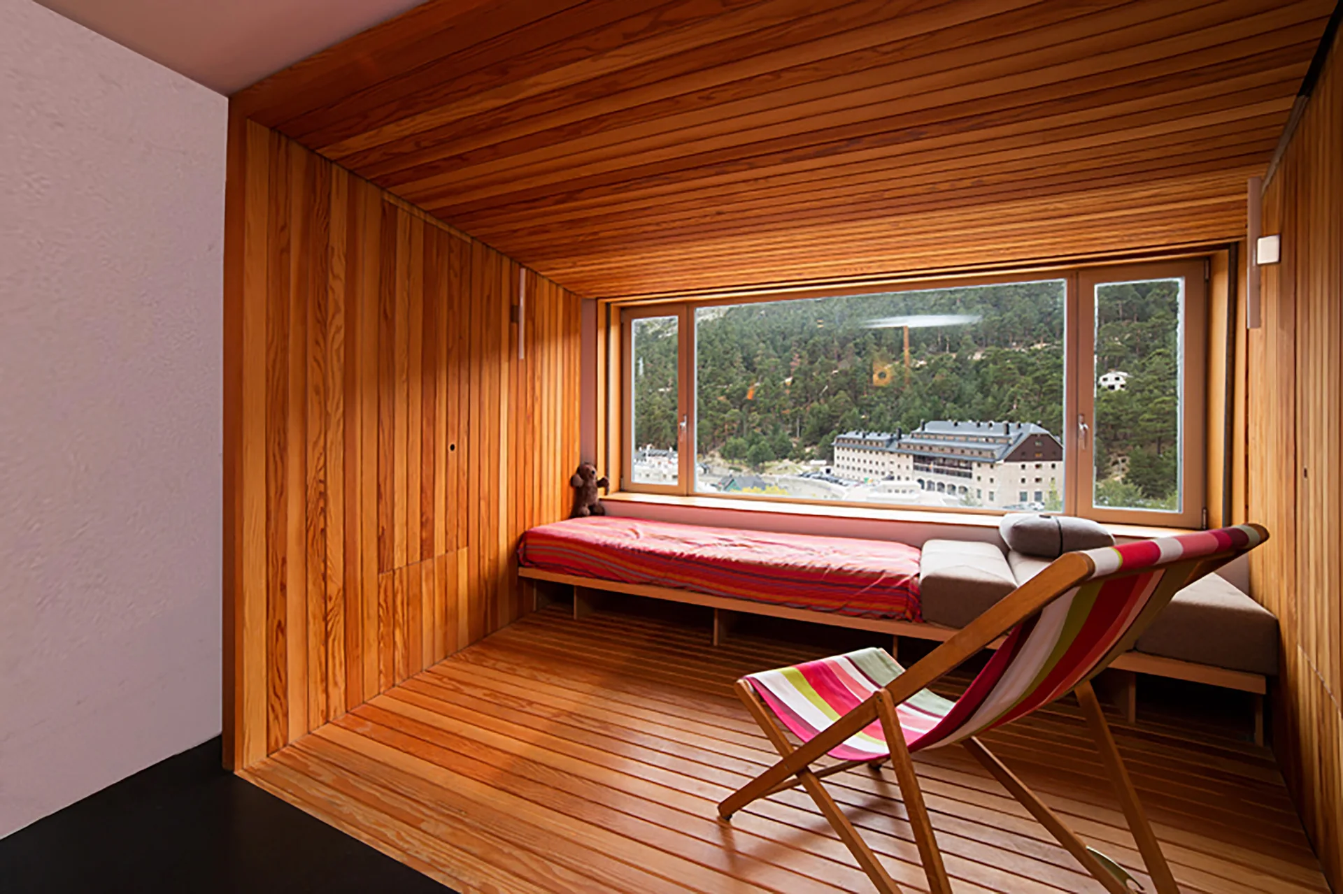 A wood-paneled cabin bedroom features a built-in daybed beneath a large window with mountain views and a colorful folding deck chair.