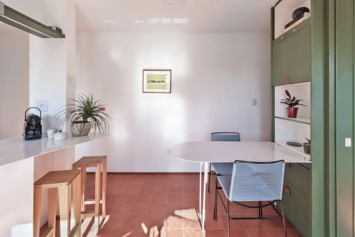 A minimalist dining area featuring a sage green built-in cabinet, an integrated white table with blue corded chairs, and terracotta floor tiles.