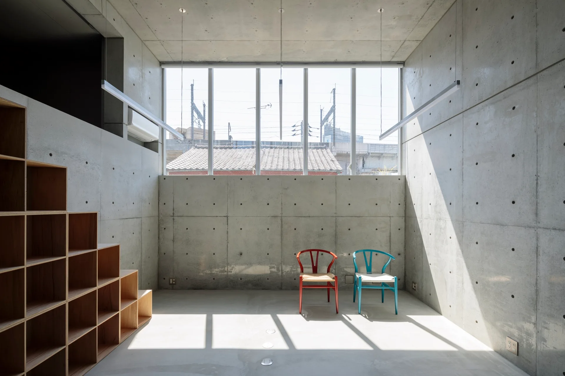 A minimalist concrete room with a stepped plywood shelving unit, a large horizontal window, and two colored chairs on the floor.
