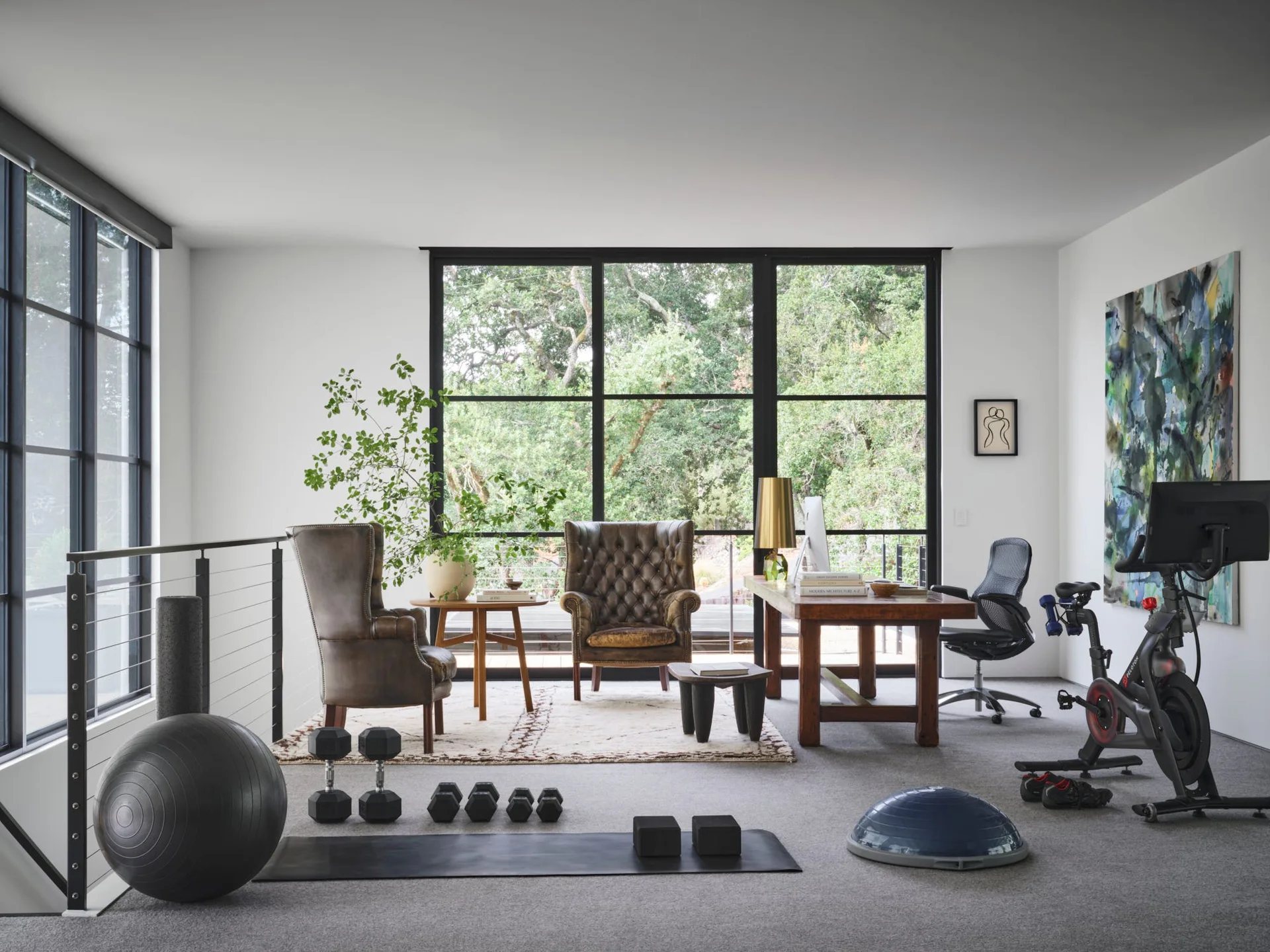 A multi-purpose home office and workout room features a wooden desk, tufted leather armchairs, and gym equipment next to large black-framed windows.