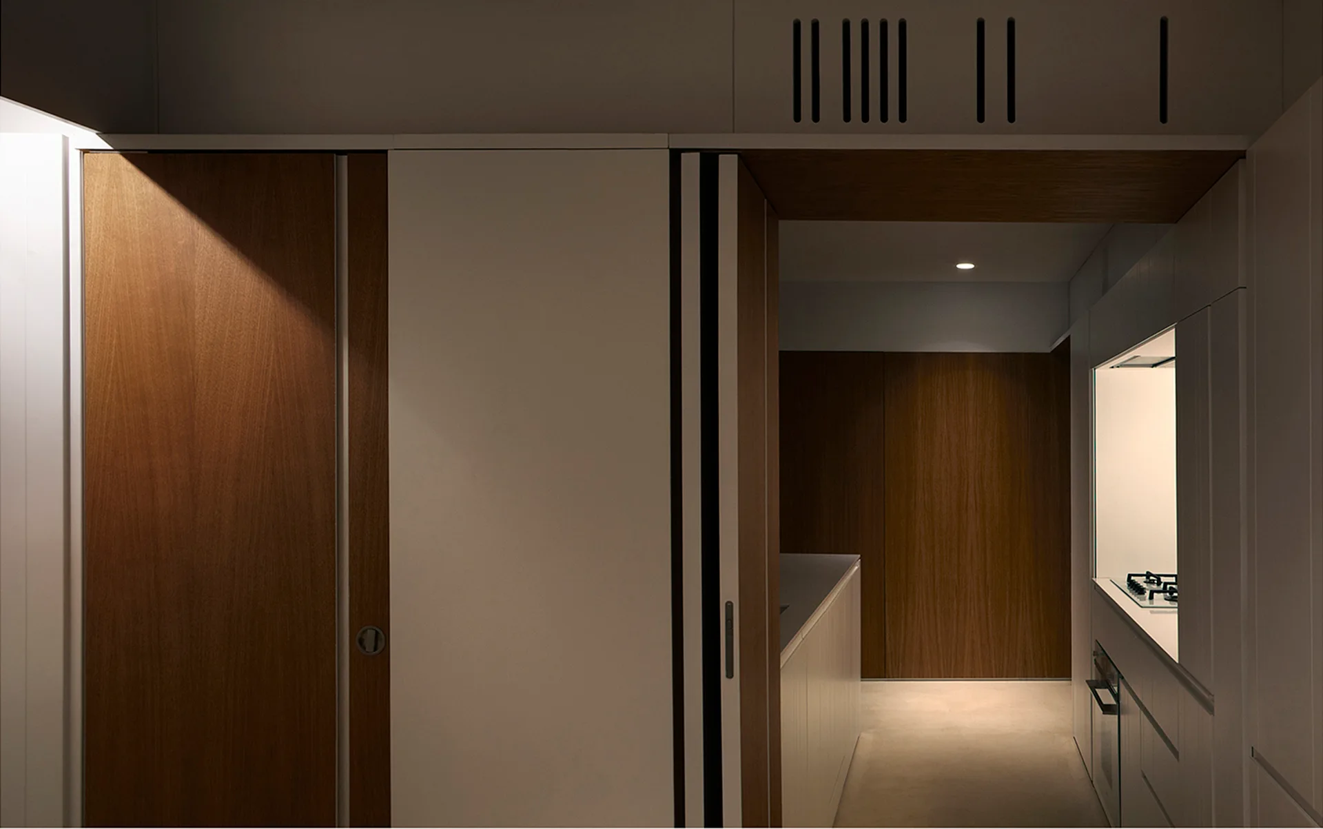 A minimalist corridor kitchen showing integrated flat-front white cabinetry alongside rich wood veneer sliding doors and a recessed stovetop niche.