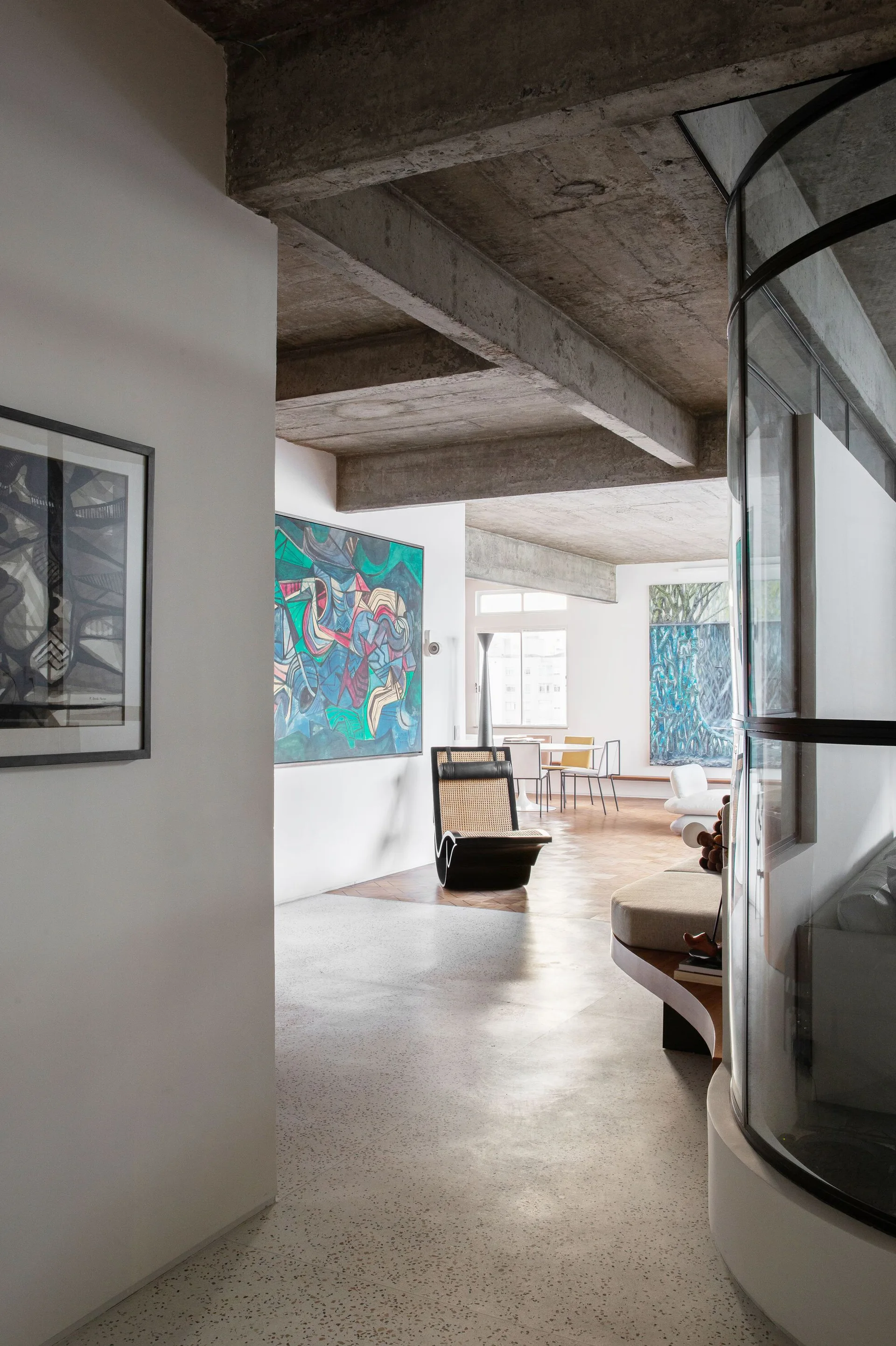 An open-concept living room features exposed concrete ceiling beams, a curved glass partition, abstract paintings, and a rattan lounge chair.