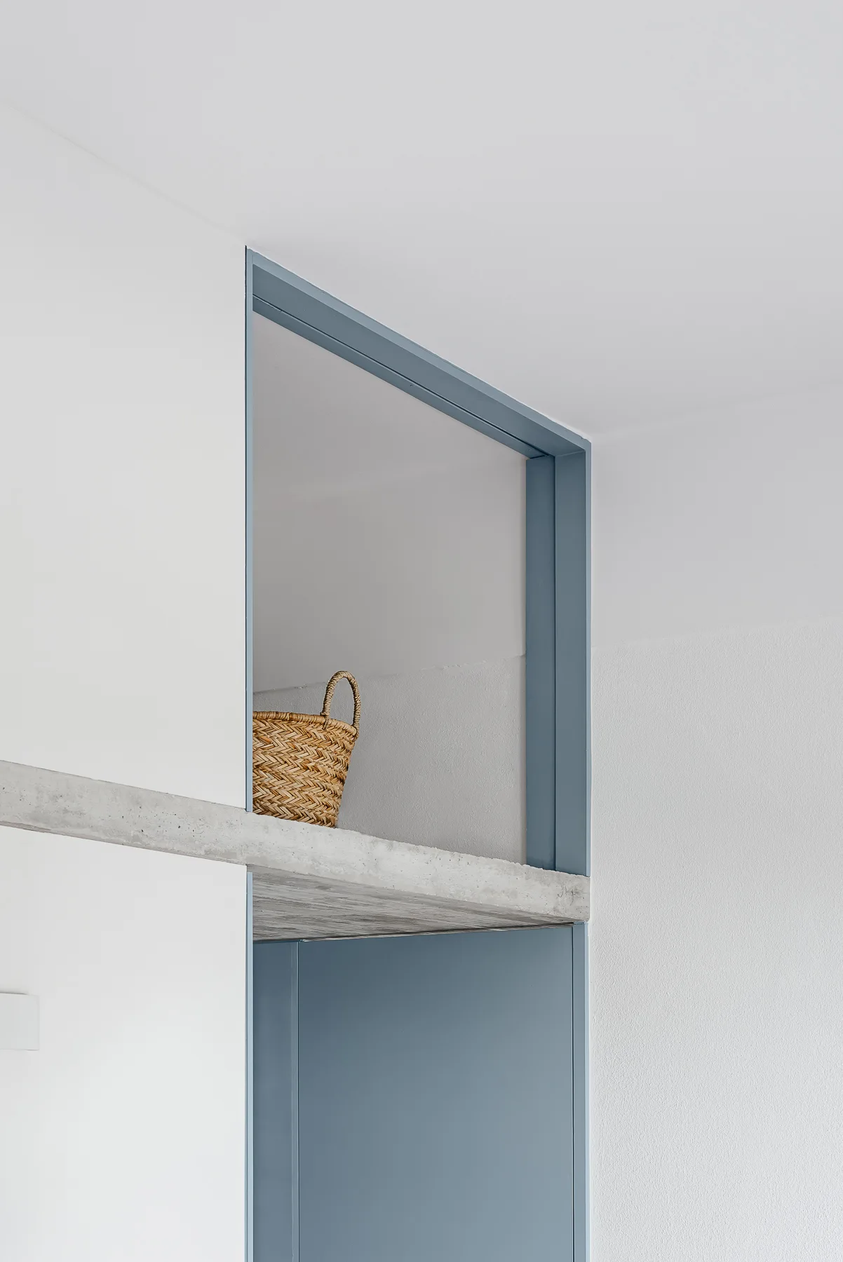 A small wicker basket sits on an exposed concrete shelf integrated into a blue painted door frame set against white walls.