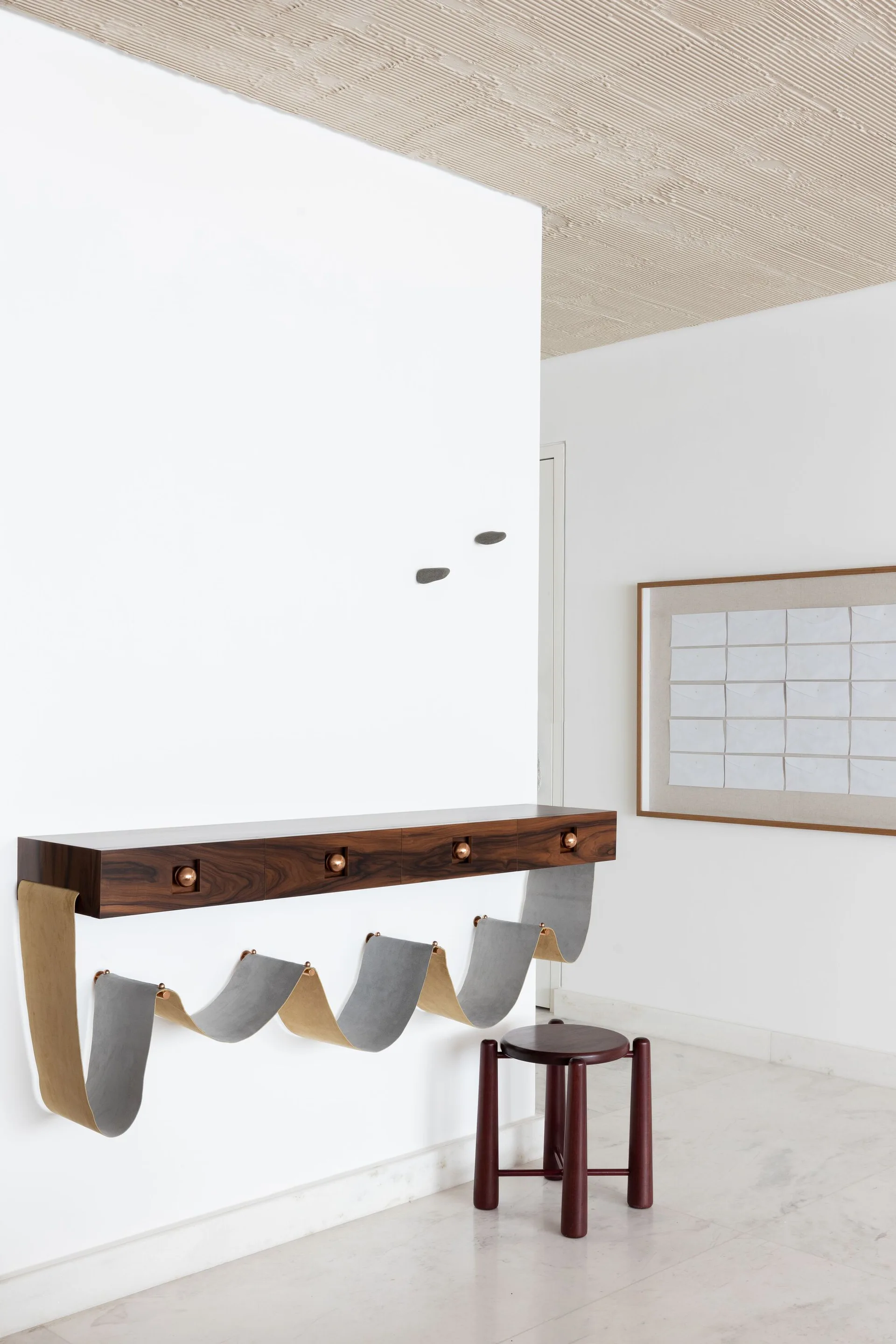A minimalist white entryway featuring a wall-mounted dark wood console with hanging fabric slings, a matching round stool, and a textured beige ceiling.
