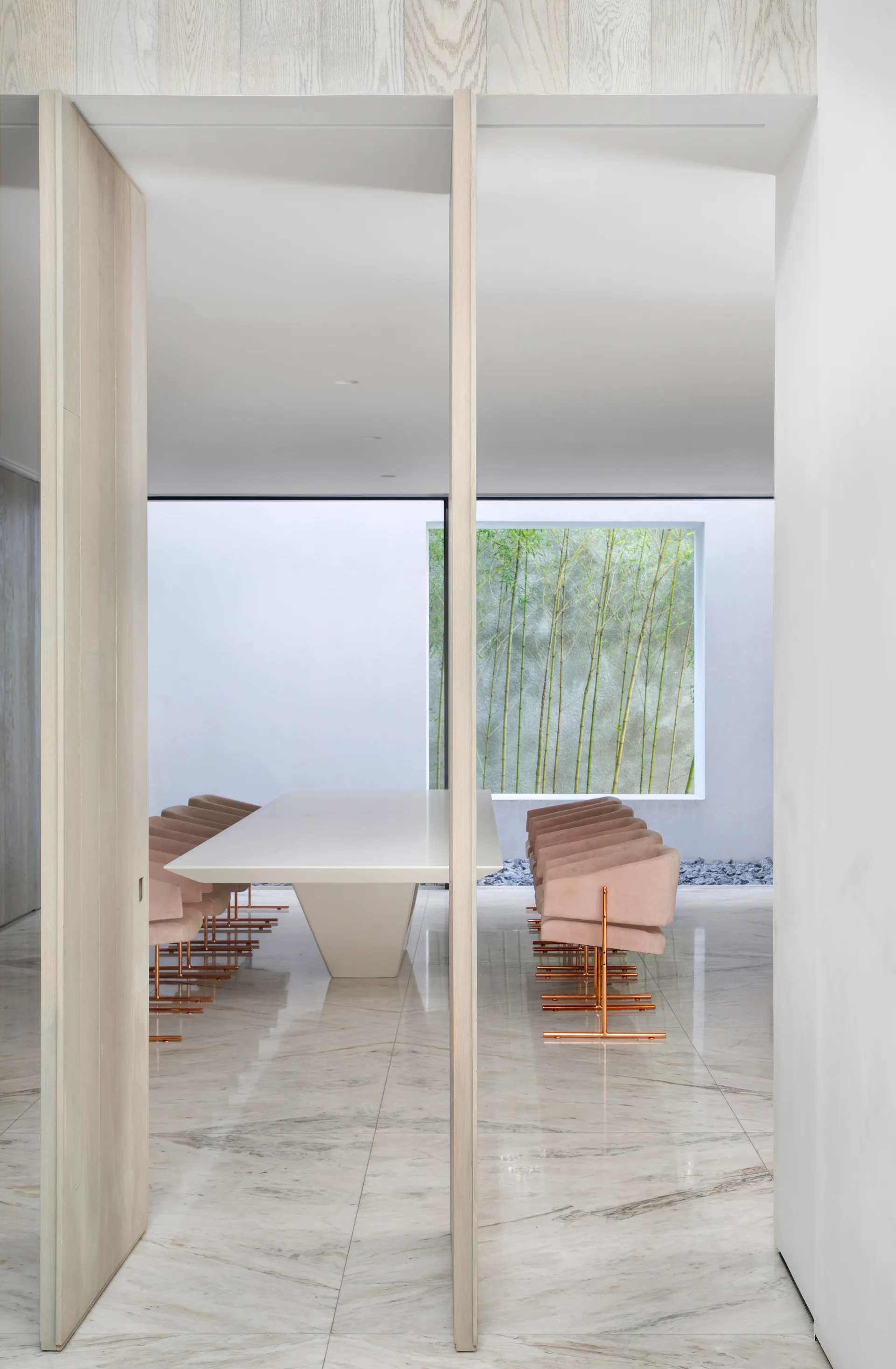 A minimalist dining room viewed through a wood doorway, featuring a white table, blush pink chairs, marble flooring, and a large window facing bamboo.
