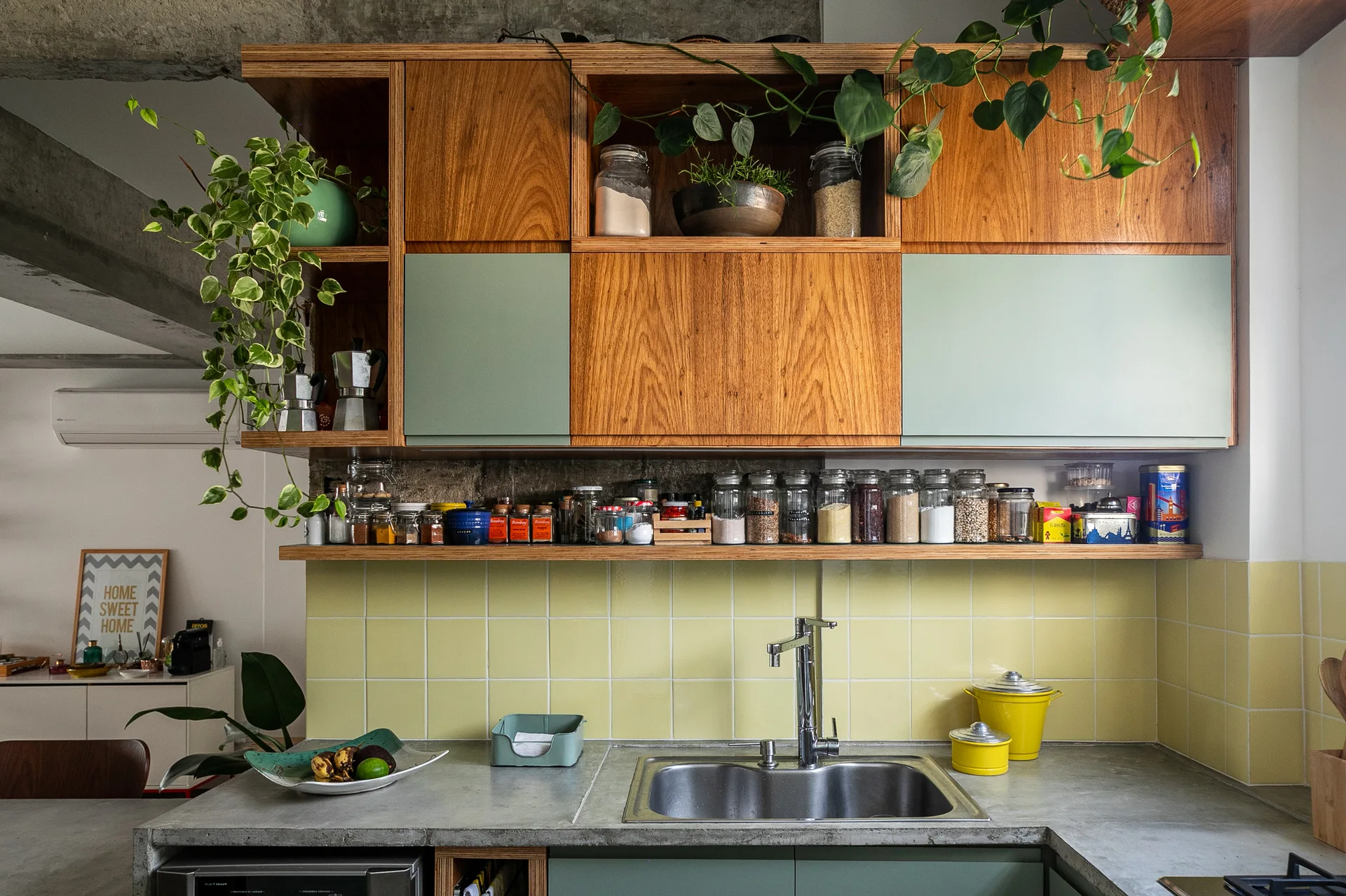 Wood and green kitchen cabinets hang above a yellow tiled backsplash, a concrete countertop, and a stainless steel double sink.