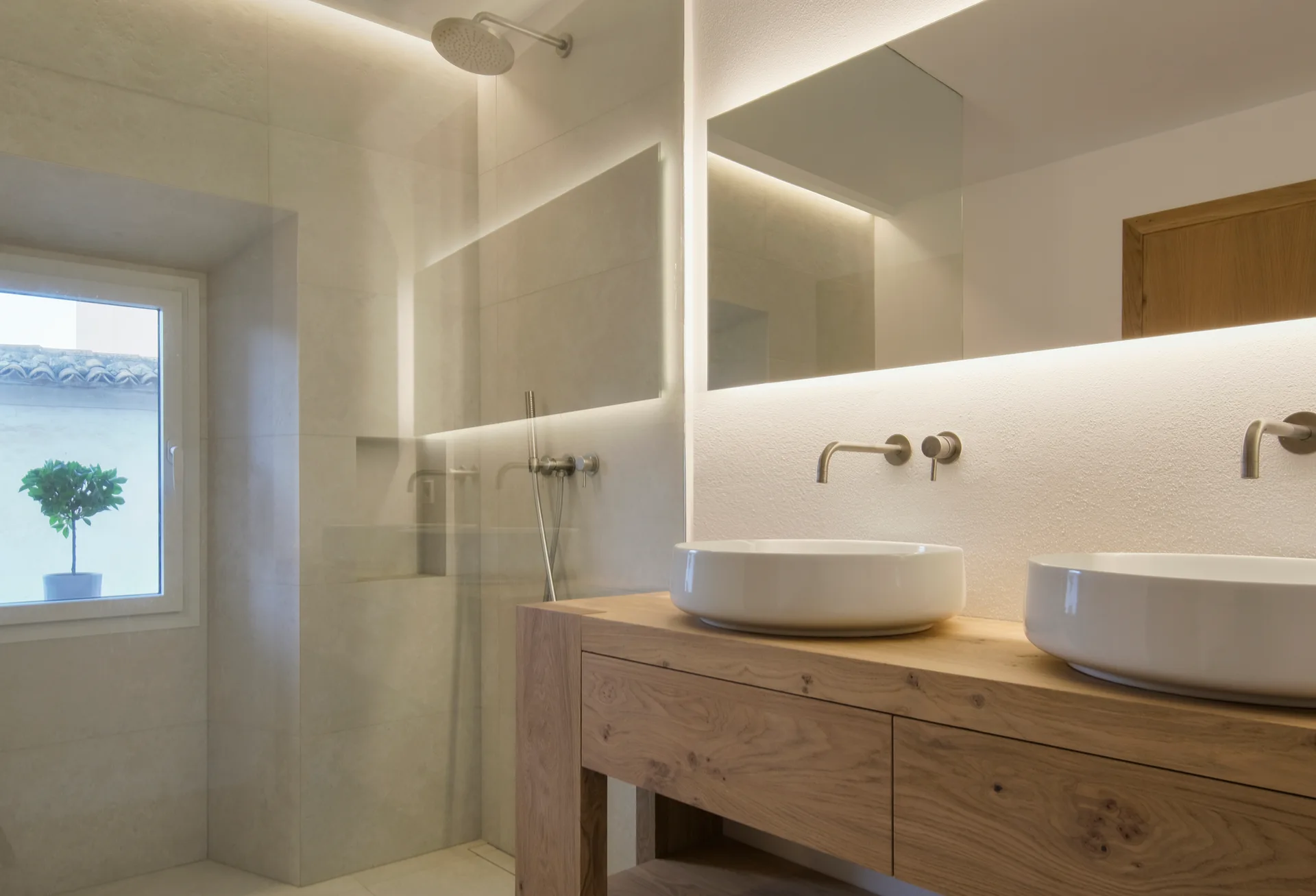 A modern bathroom showing a light wood double vanity with two white ceramic vessel sinks and backlit rectangular mirrors.