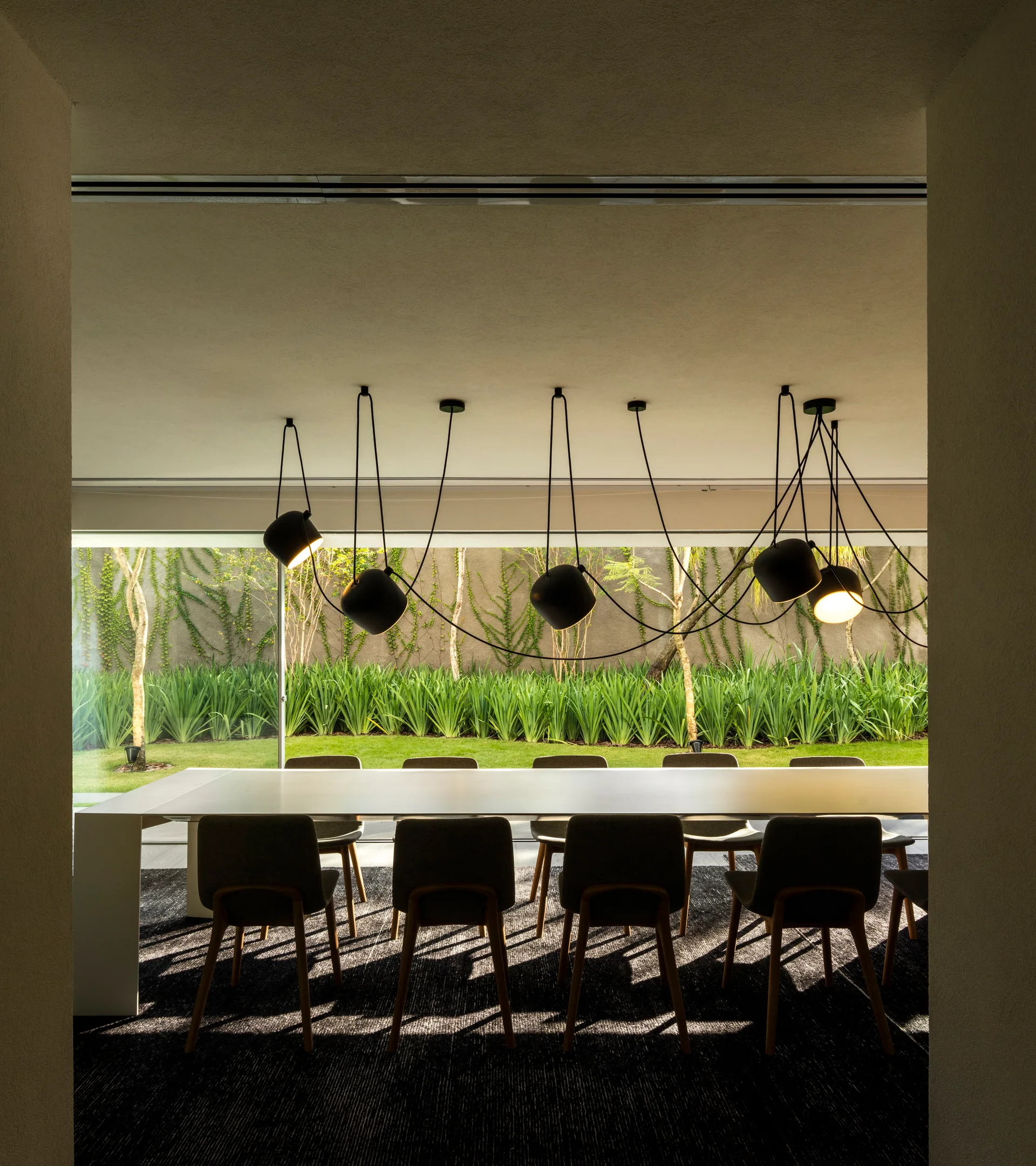 A long white dining table with chairs sits on a dark rug before a wall of glass looking out to a lush green garden with pendant lights above.
