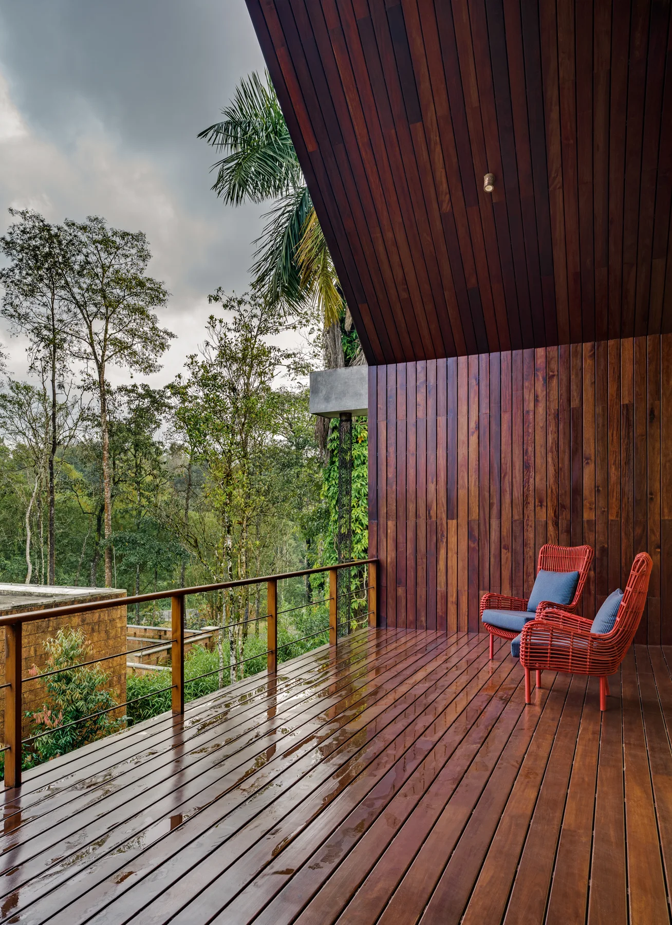 A covered wooden terrace with dark vertical wood cladding, a wet polished deck, and two red woven armchairs overlooking a dense green forest.