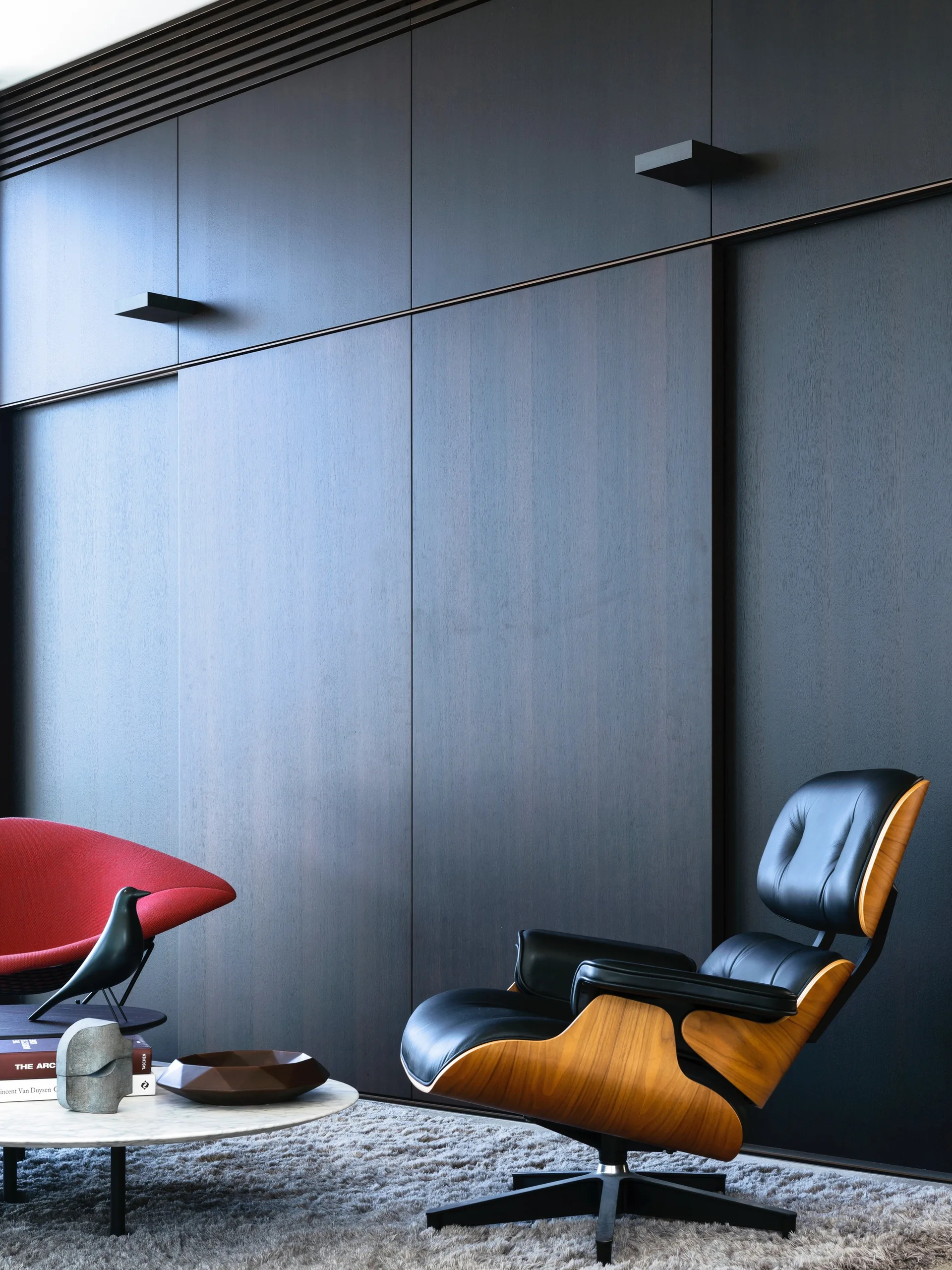 A modern room features dark wood wall panels, a black leather and plywood lounge chair, a red accent chair, and a low stone coffee table on a grey rug.