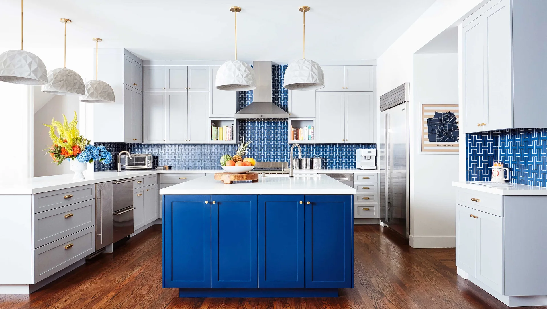 A modern kitchen featuring a bright blue central island, light grey cabinets, a blue geometric patterned backsplash, and white dome pendant lights.