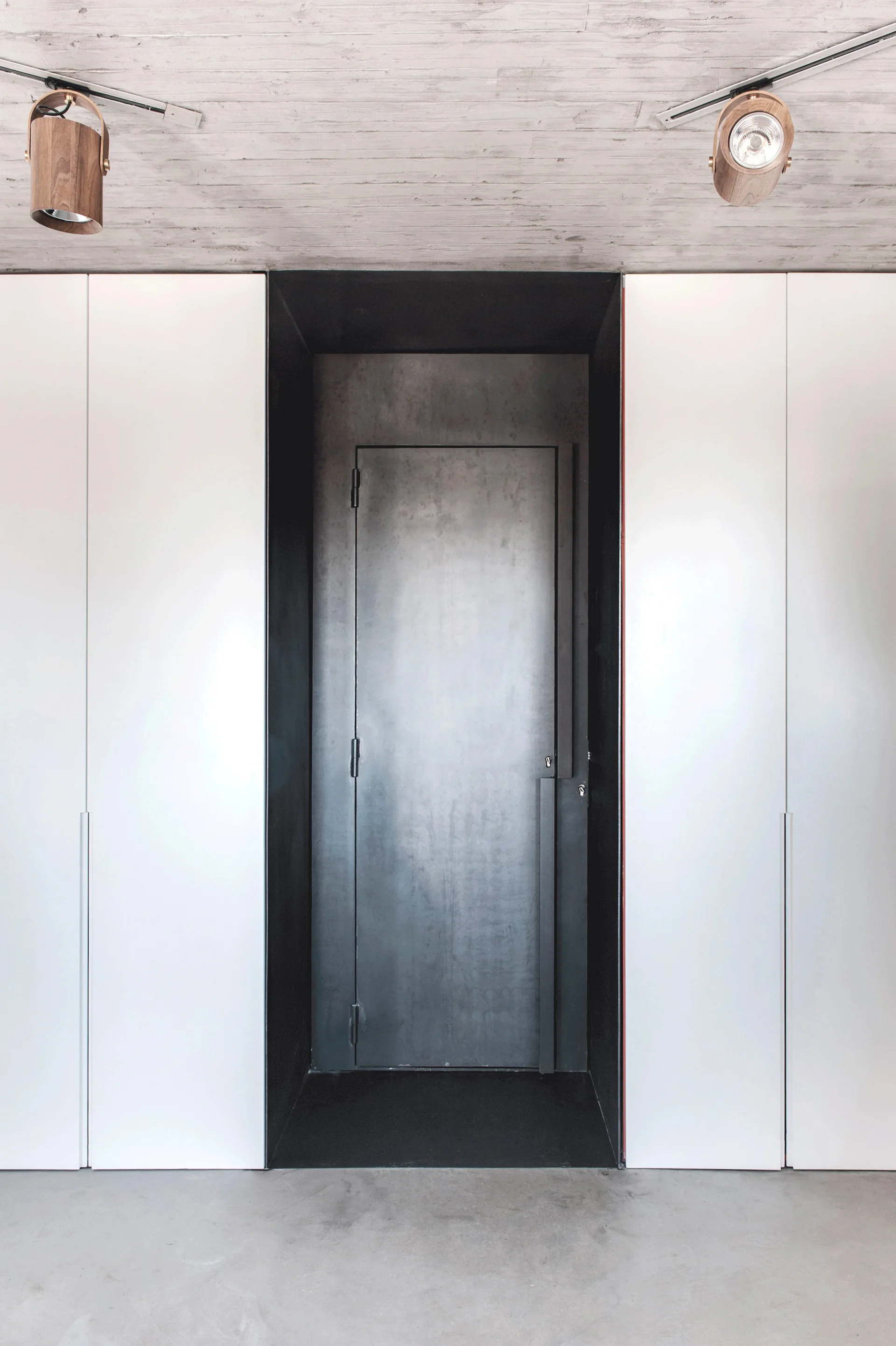 A minimalist hallway featuring a central dark metal door set in a black recess, flanked by white full-height cabinets under a concrete ceiling.