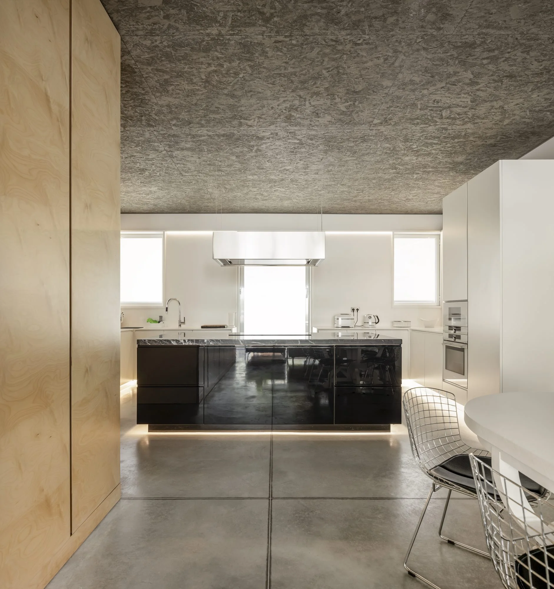 A minimalist kitchen features a glossy black island with under-cabinet lighting, a textured OSB ceiling, concrete floors, and a light wood cabinet.
