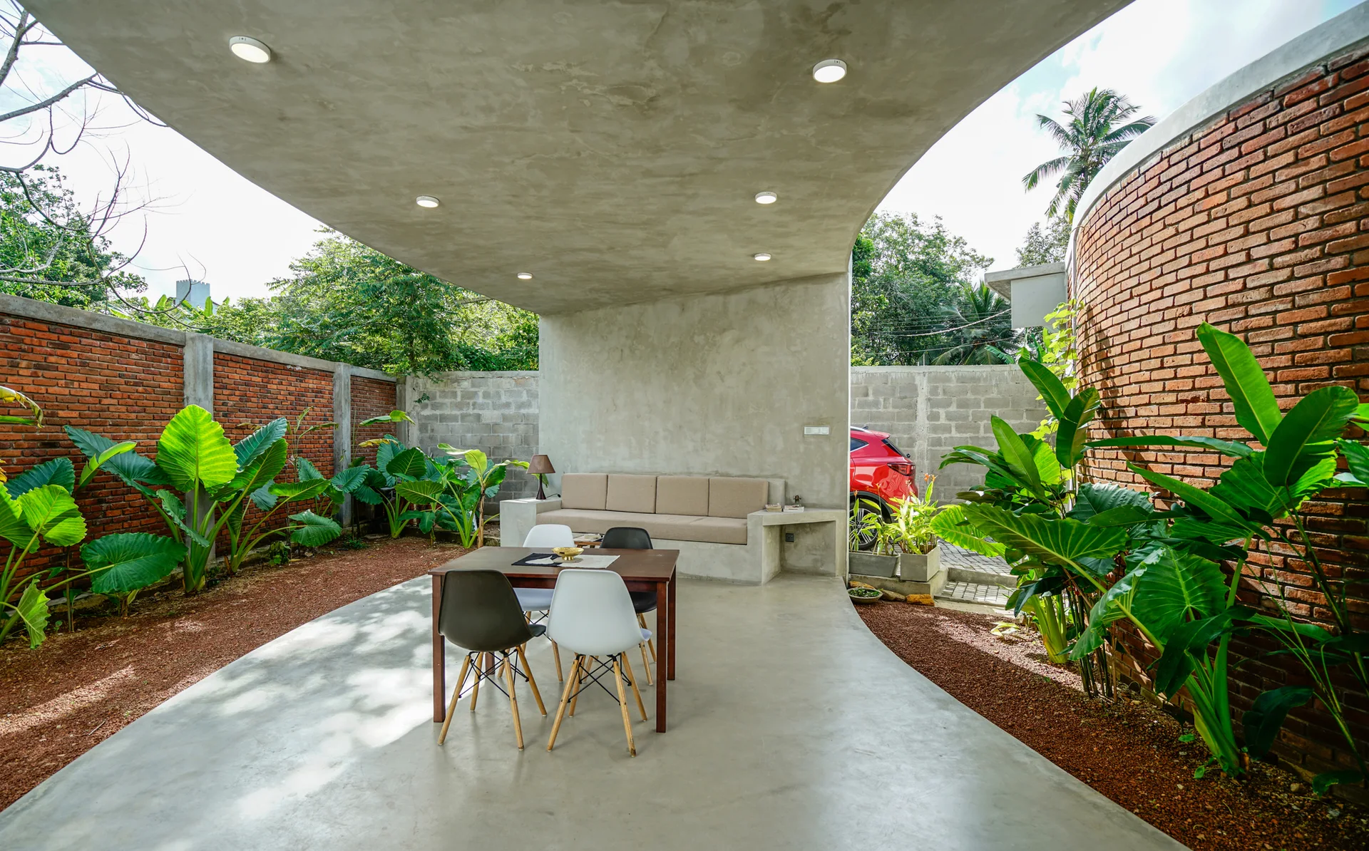 A covered concrete patio features a built-in bench, a wooden dining table with black and white chairs, and exposed red brick walls with lush green plants.