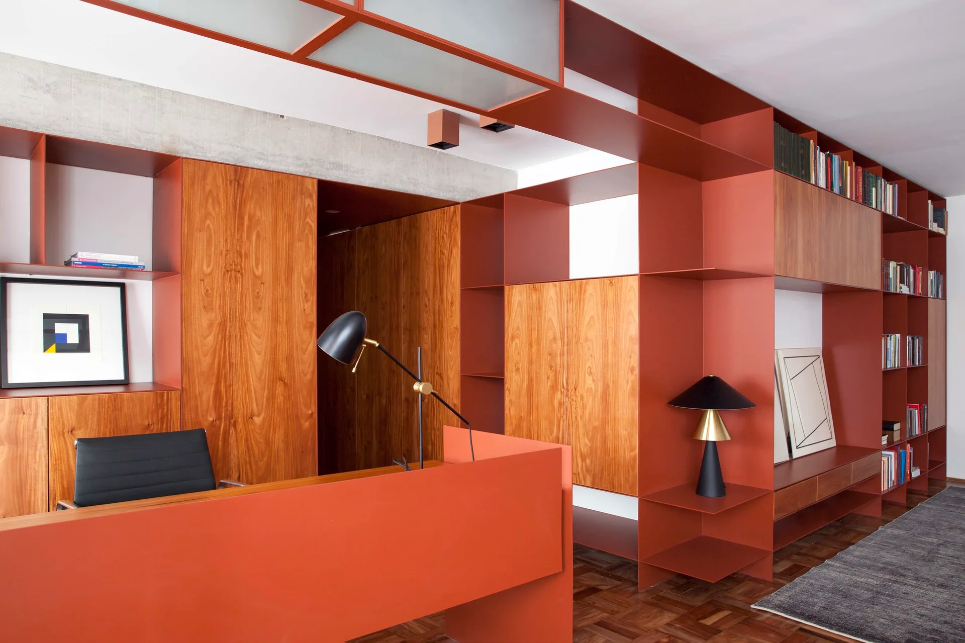 A modern home office featuring a terracotta orange metal shelving unit integrated with wood cabinets, a matching orange desk, and a black task chair.