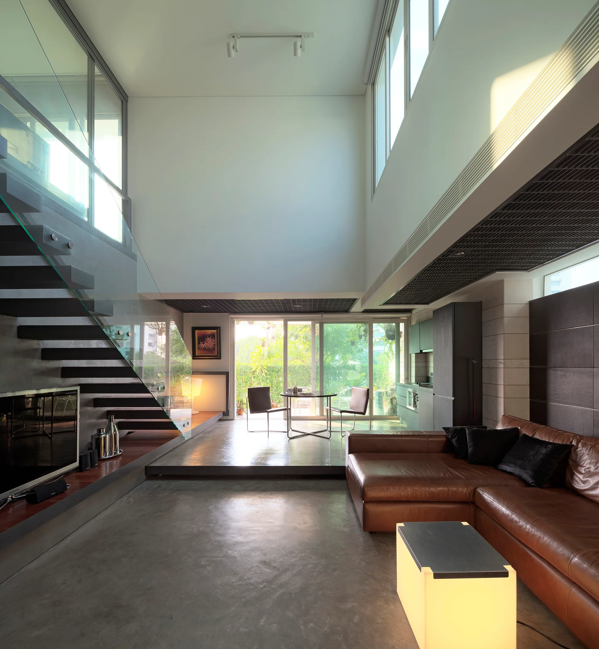 A spacious double-height living room featuring a brown leather sectional sofa, polished concrete floors, and a floating staircase with a glass railing.