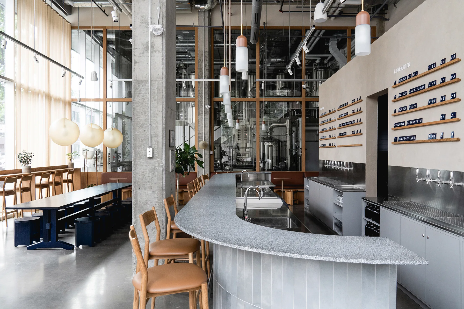 A modern brewery taproom featuring a curved grey bar counter with wooden stools and large brewing tanks behind a glass partition.