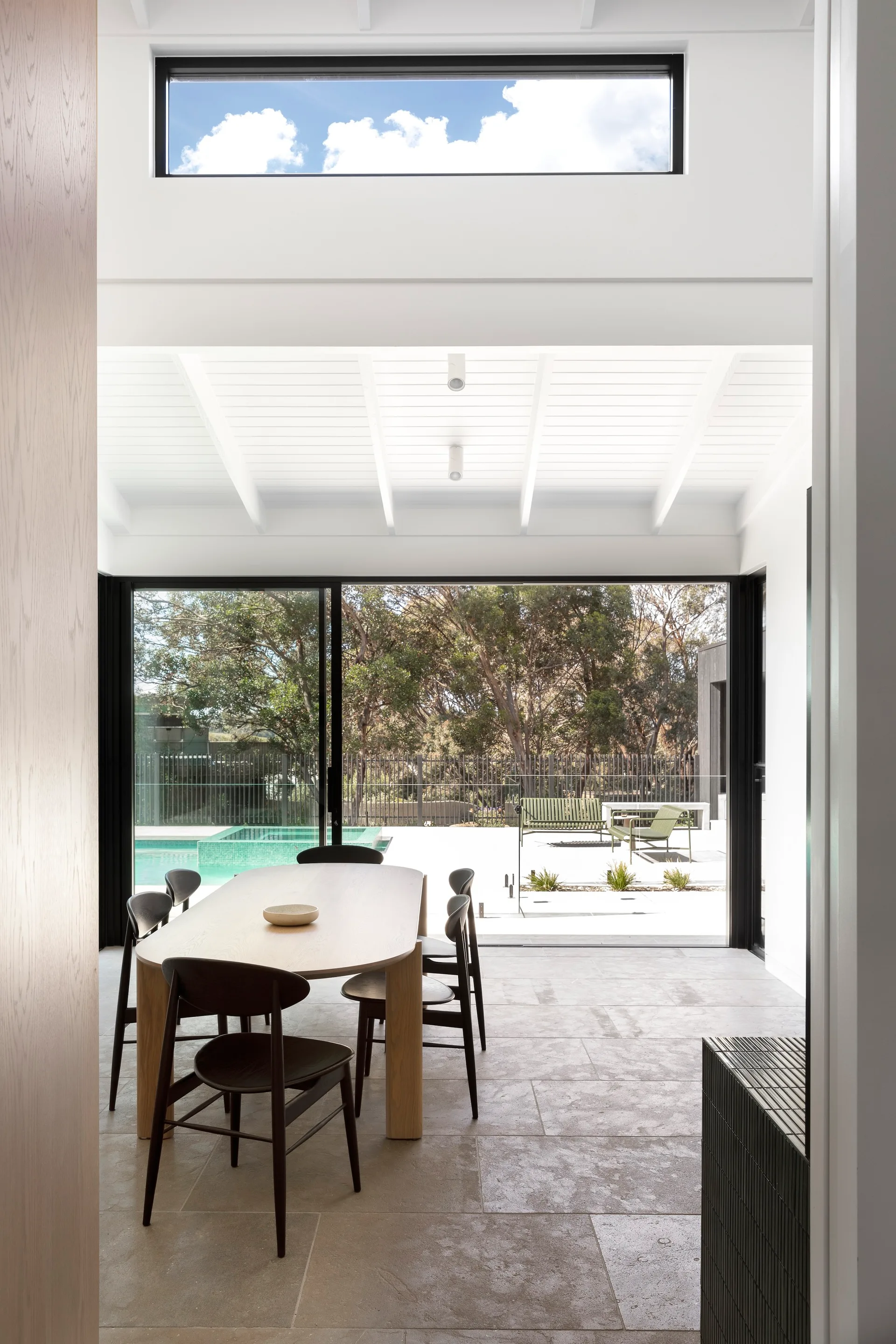 A light-filled dining room features a light wood table, dark chairs, and large sliding glass doors opening to a patio and pool.
