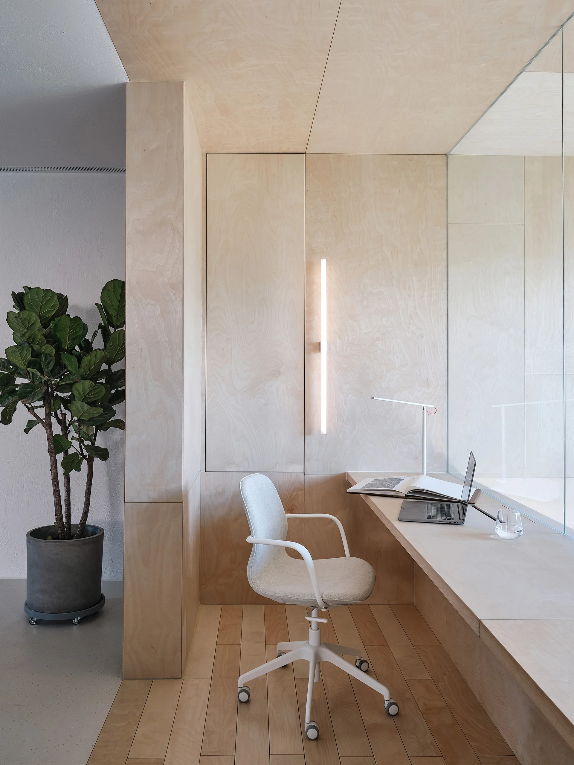 A minimalist home office nook features light wood plywood paneling, a built-in desk, a white swivel chair, and an integrated vertical wall light.