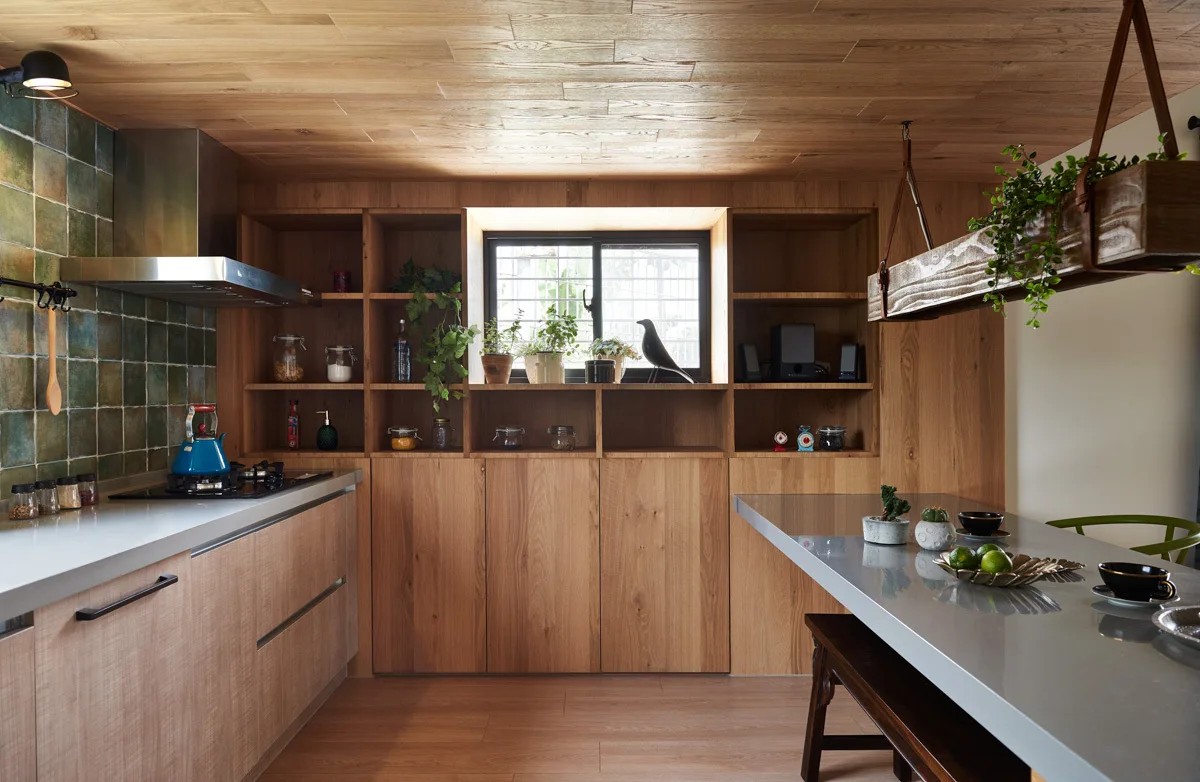 A contemporary kitchen featuring wood paneled cabinets and ceiling, a green square tile backsplash, a gray island, and a window framed by open shelves.