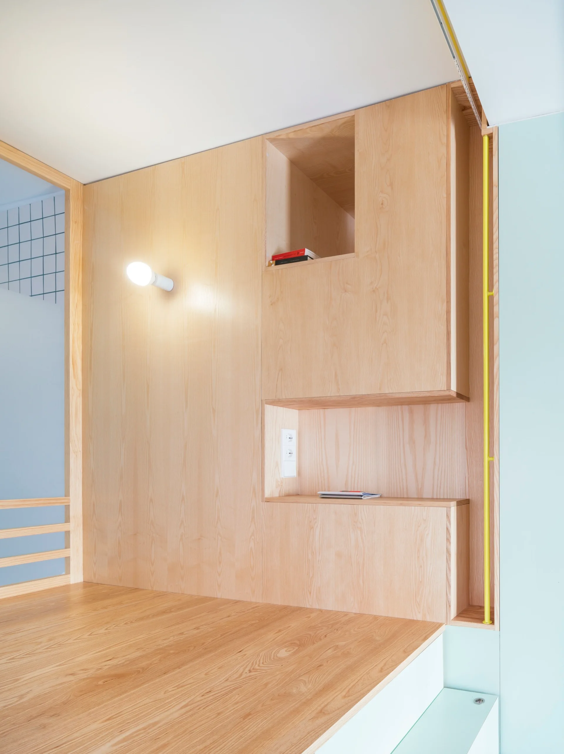 A custom light wood paneled wall features built-in storage niches, a glowing white globe sconce, and a yellow vertical metal pipe.