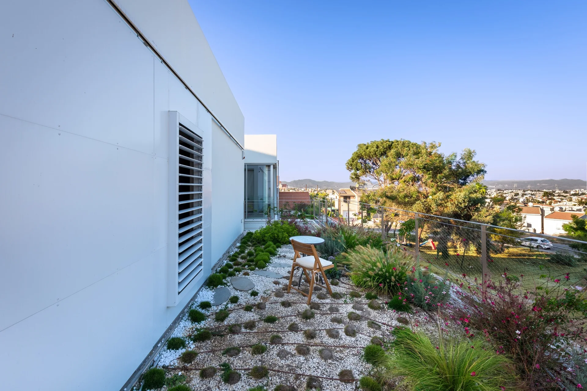 A modern white rooftop terrace features a gravel garden with tufts of grass, a metal railing, and a small wooden folding chair and bistro table.