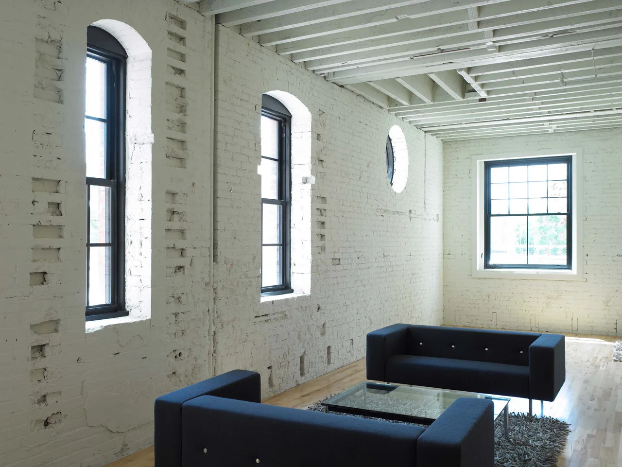 An industrial loft living room with white-painted brick walls, arched black-framed windows, exposed ceiling beams, and dark blue sofas.