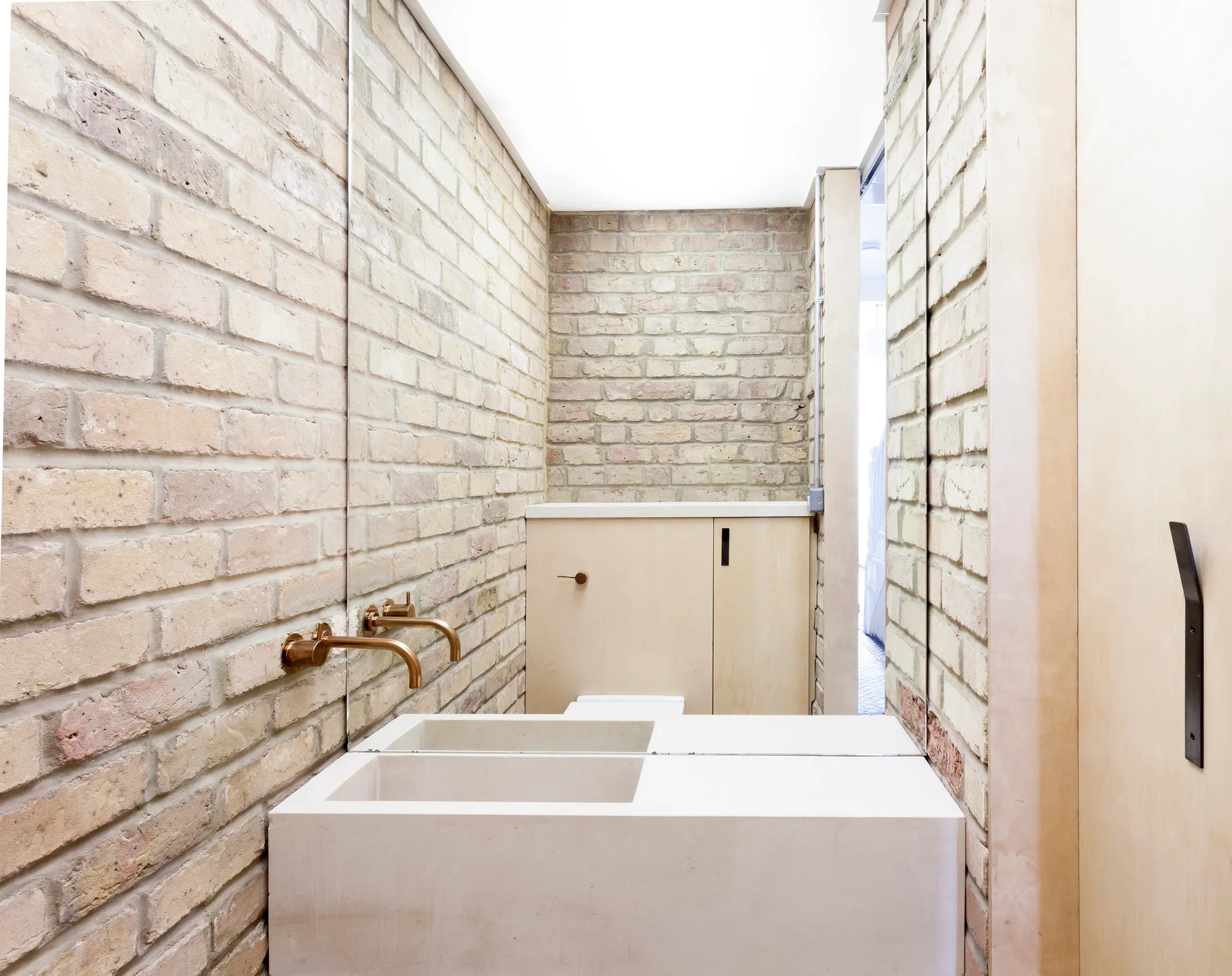 A minimalist bathroom featuring light-colored exposed brick walls, a monolithic white stone sink, and a wall-mounted copper faucet on a large mirror.