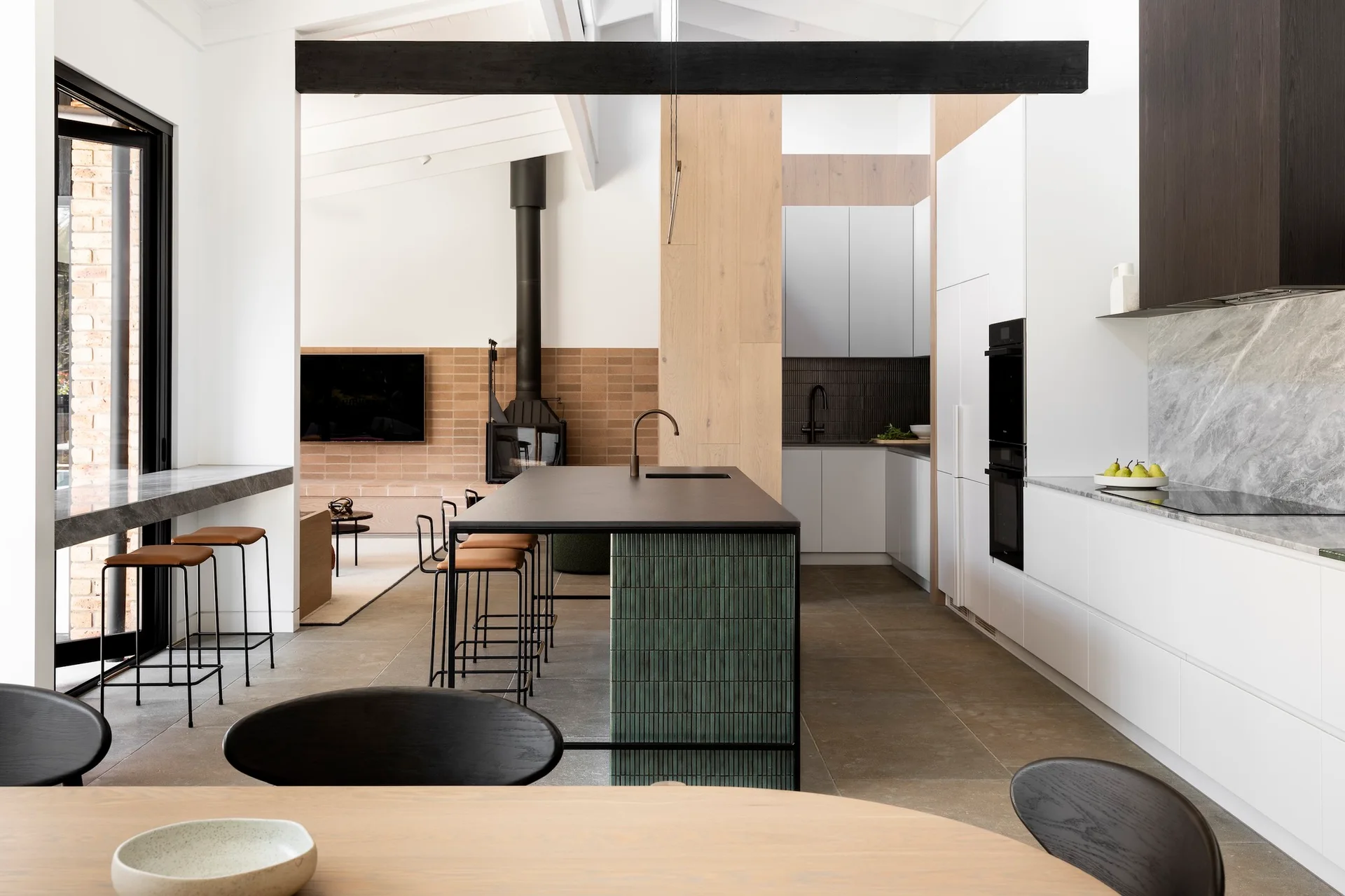 A contemporary open-concept kitchen featuring a dark-topped island with vertical green tiles, minimalist white cabinetry, and a black ceiling beam.