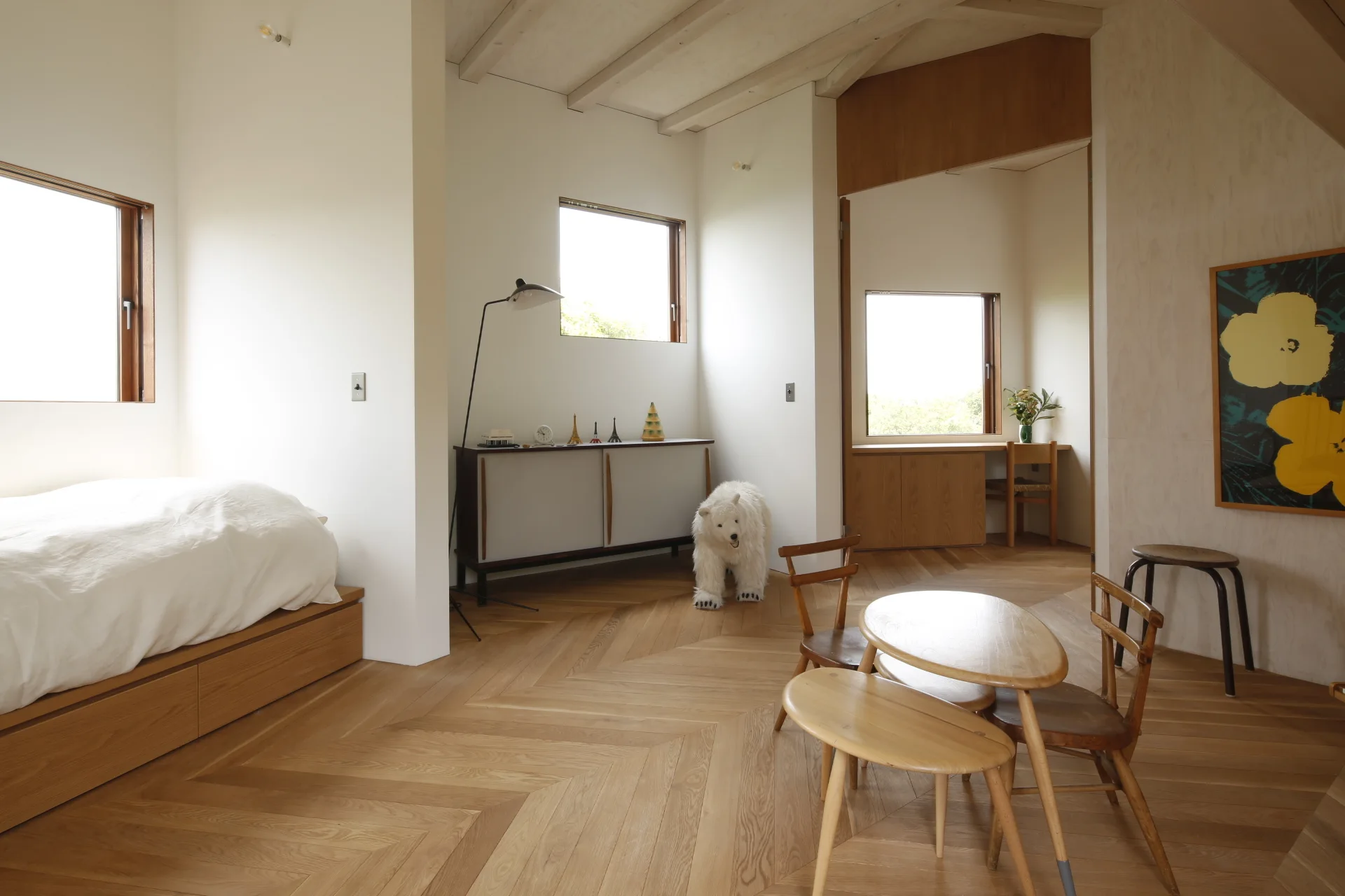 A minimalist white bedroom featuring light wood herringbone flooring, exposed ceiling beams, a low platform bed, and small wooden furniture pieces.