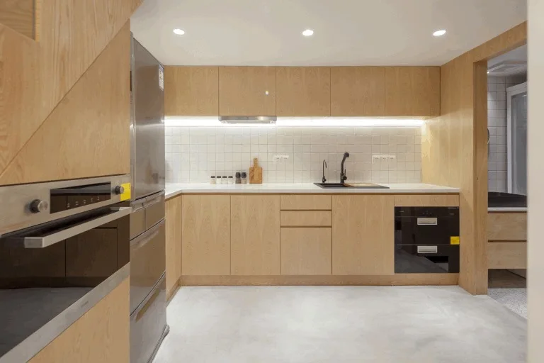 A modern kitchen with light wood flat-front cabinetry, white square tile backsplash with under-cabinet lighting, and integrated appliances.