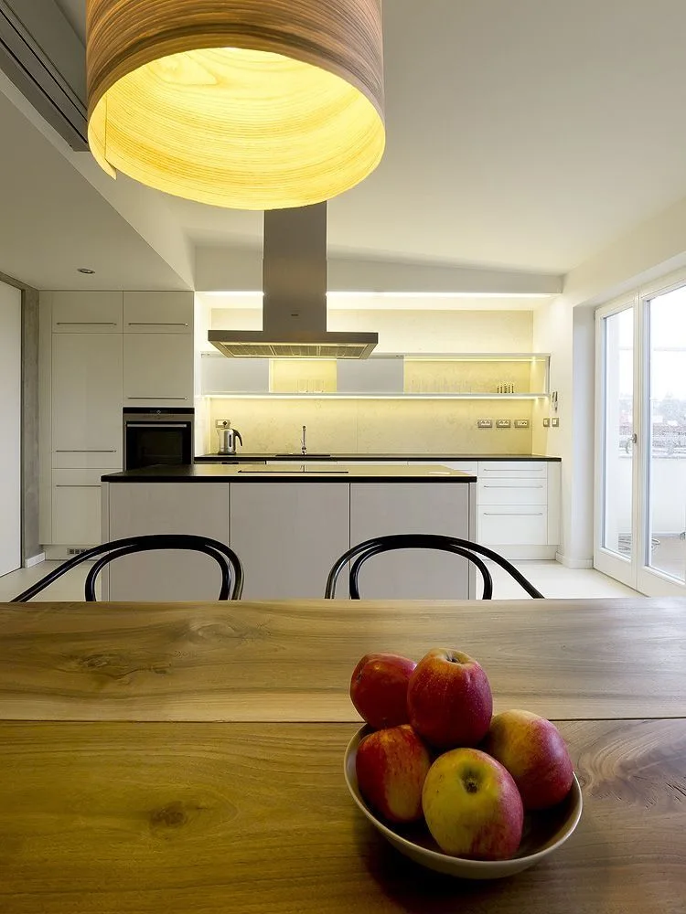 A minimalist white kitchen features a large wood-grained pendant light hanging above a wood dining table with a bowl of apples in the foreground.